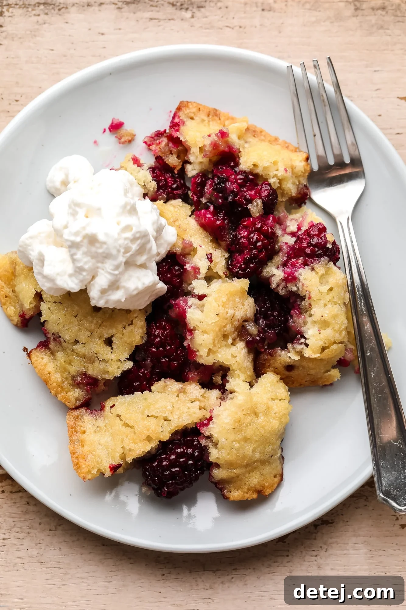 A warm scoop of Vegan Blackberry Cobbler on a white plate, garnished with vegan whipped cream and a fork on the side. This easy summer dessert is perfect with ice cream.
