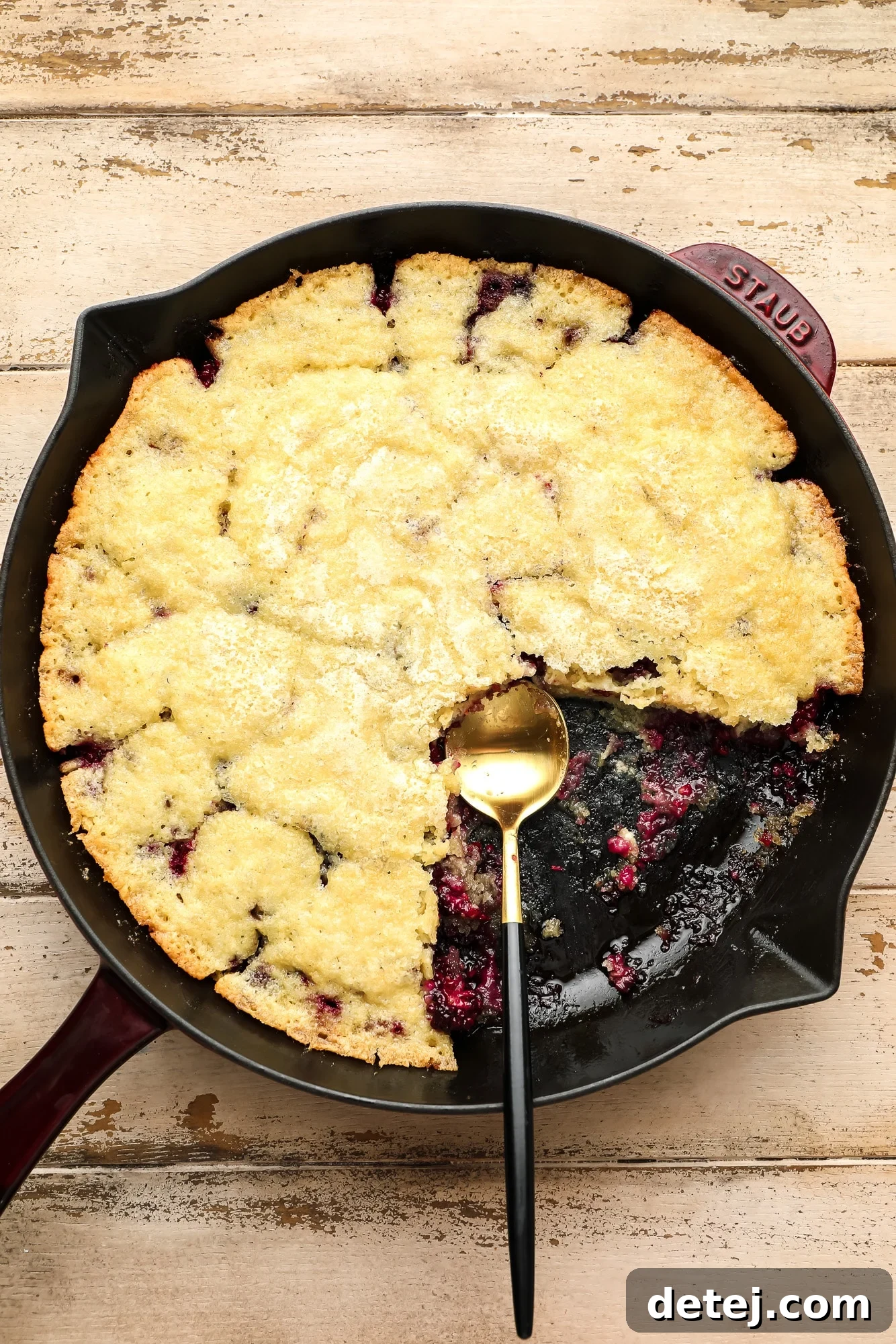 A serving spoon lifting a generous portion of baked blackberry cobbler from a black cast iron skillet, showing the golden crust and bubbling fruit underneath.