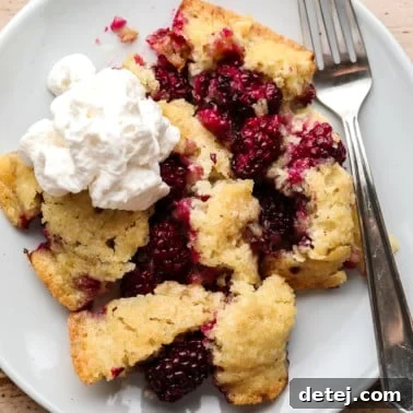 A scoop of Vegan Blackberry Cobbler on a white plate with a dollop of vegan whipped cream on top and a fork on the side.