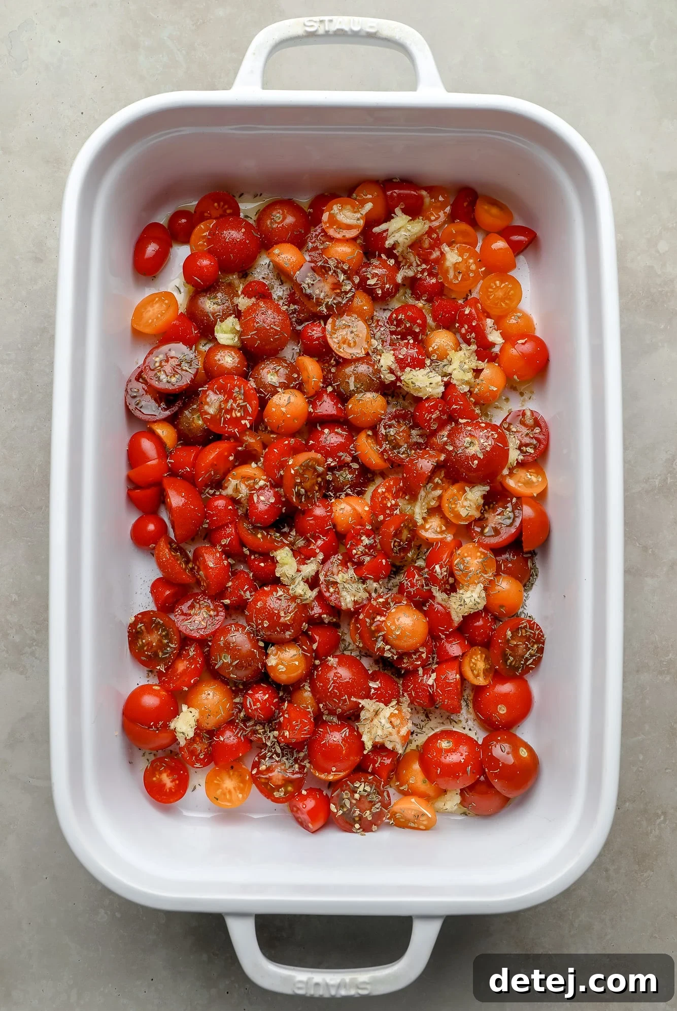 Sliced cherry tomatoes and minced garlic, seasoned with dried oregano and thyme, neatly arranged in a white baking dish before going into the oven.