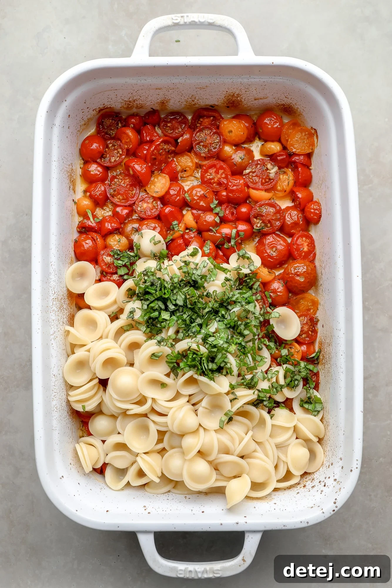 Cooked orecchiette pasta and a generous amount of fresh basil leaves are added on top of the warm roasted cherry tomatoes in a white baking dish, ready for tossing.