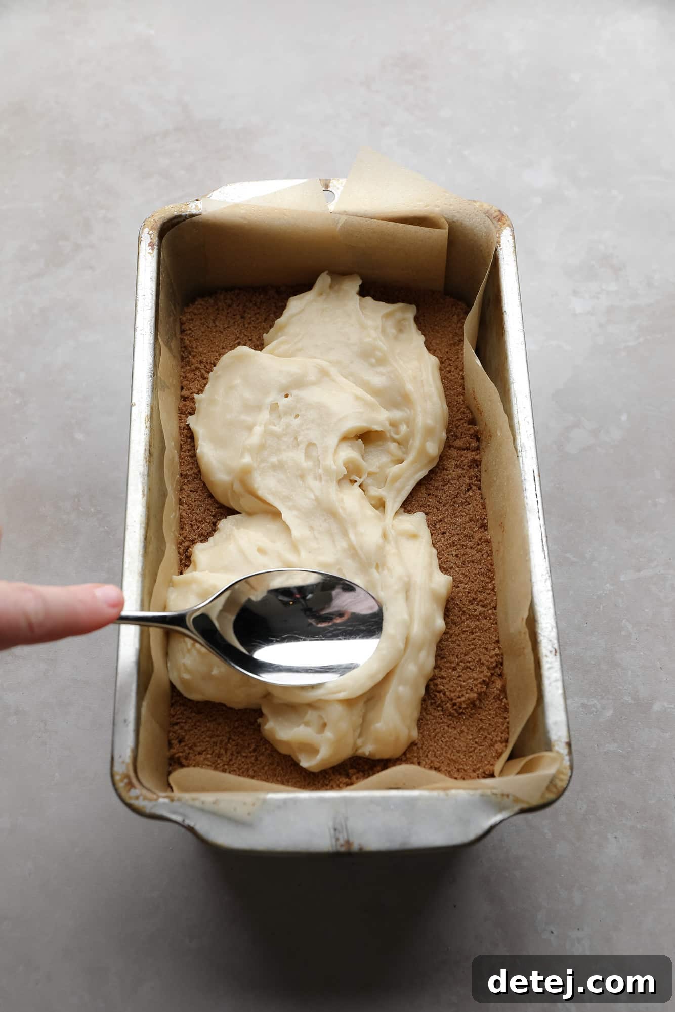 Using a spoon to spread the second layer of bread batter on top of a cinnamon sugar layer in a loaf pan.