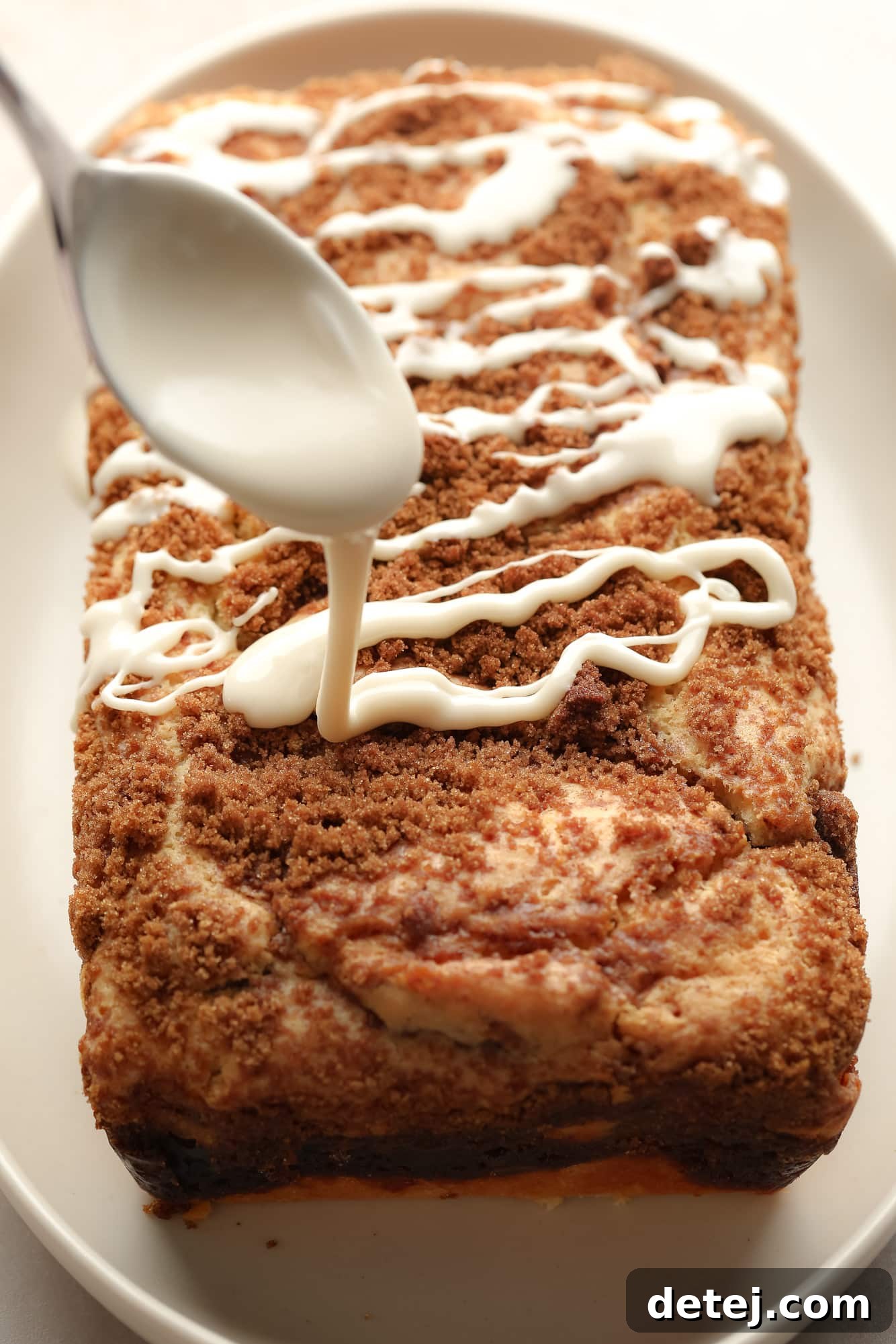Drizzling a white vanilla glaze from a spoon on top of a cooled loaf of vegan cinnamon swirl bread.
