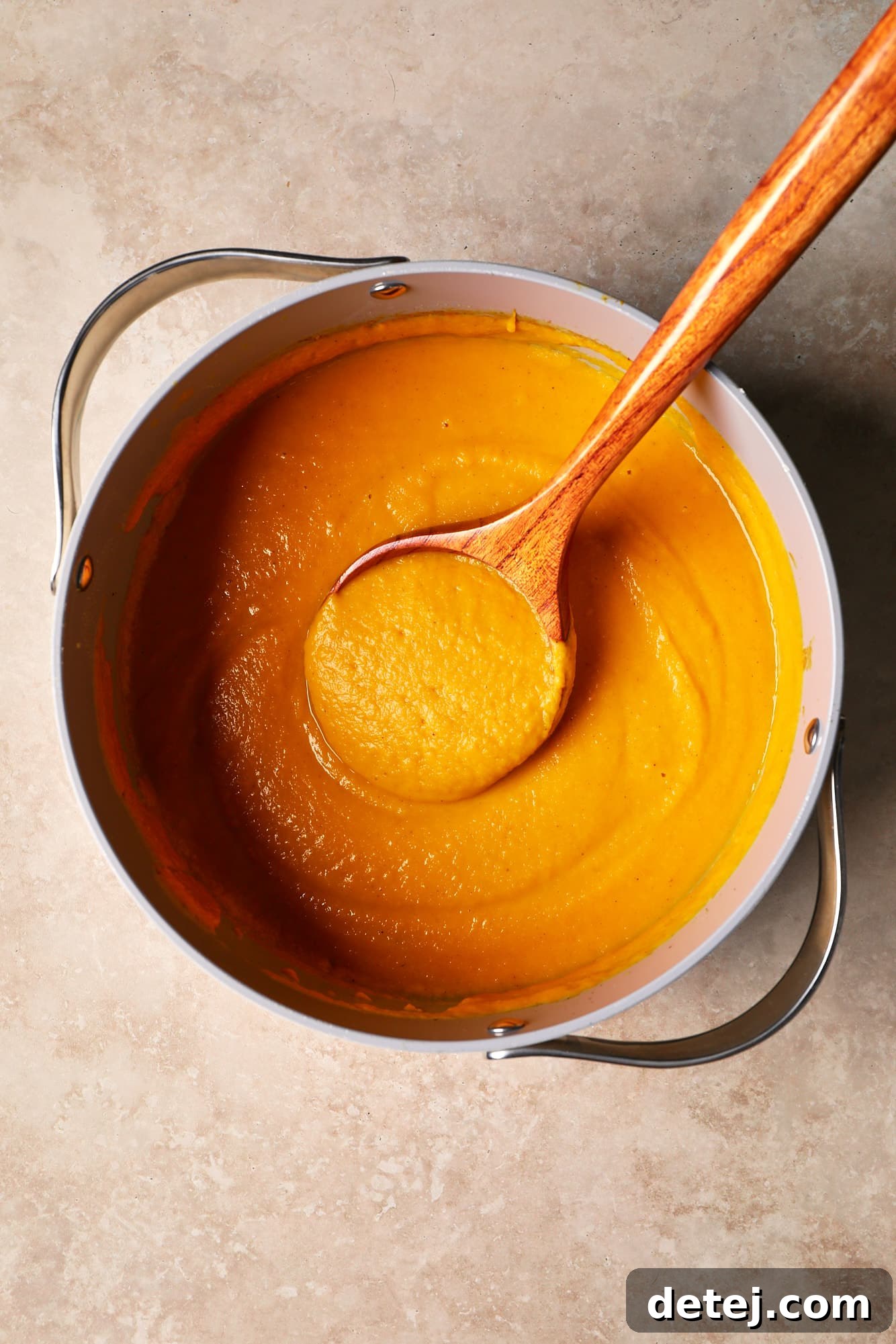 A rustic wooden spoon resting gently in a large pot filled with rich, golden Butternut Squash and Sweet Potato Soup, highlighting its smooth, creamy texture.