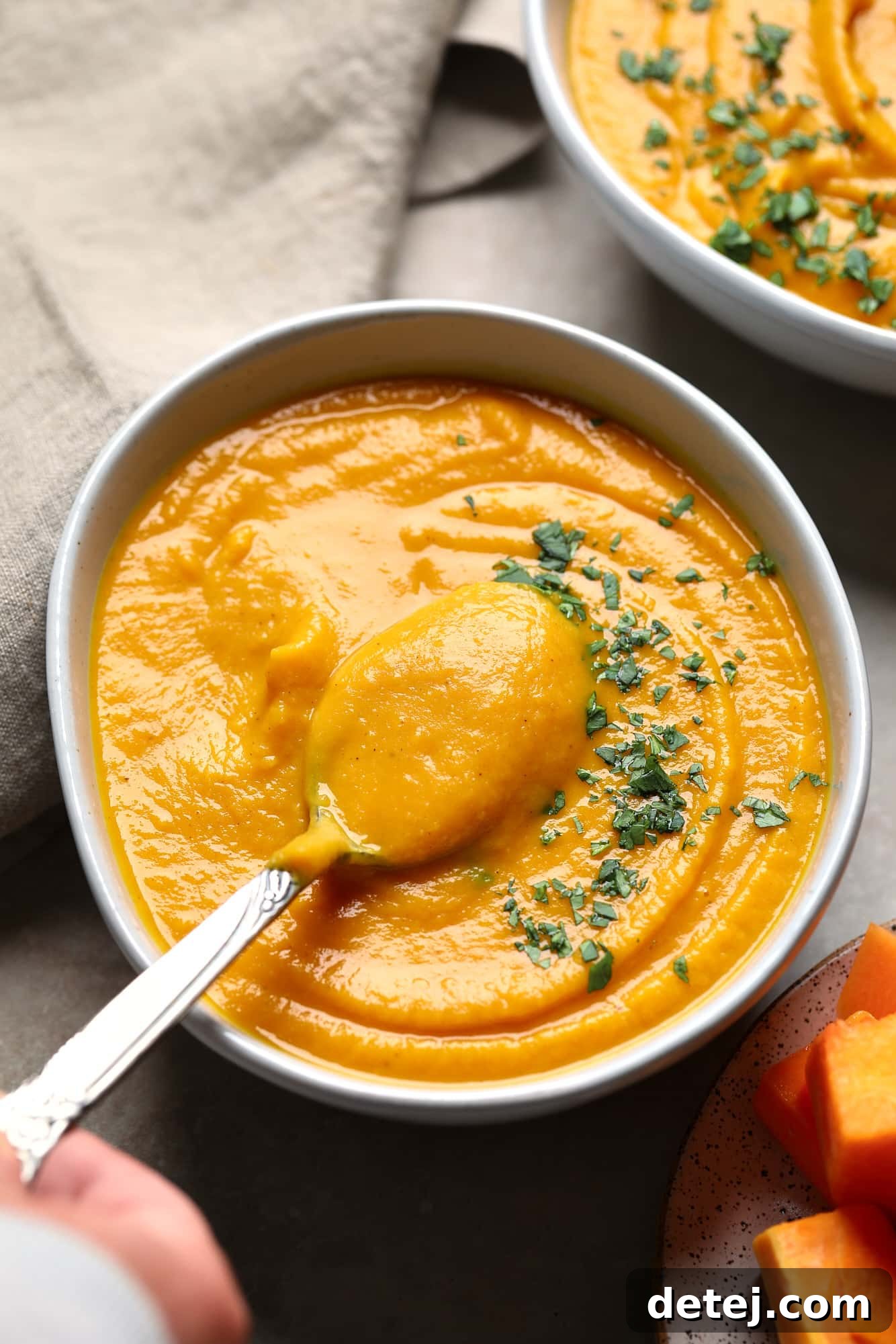 A hand gently lifting a spoon from a bowl of luscious Butternut Squash and Sweet Potato Soup, showcasing its creamy texture and inviting warmth.