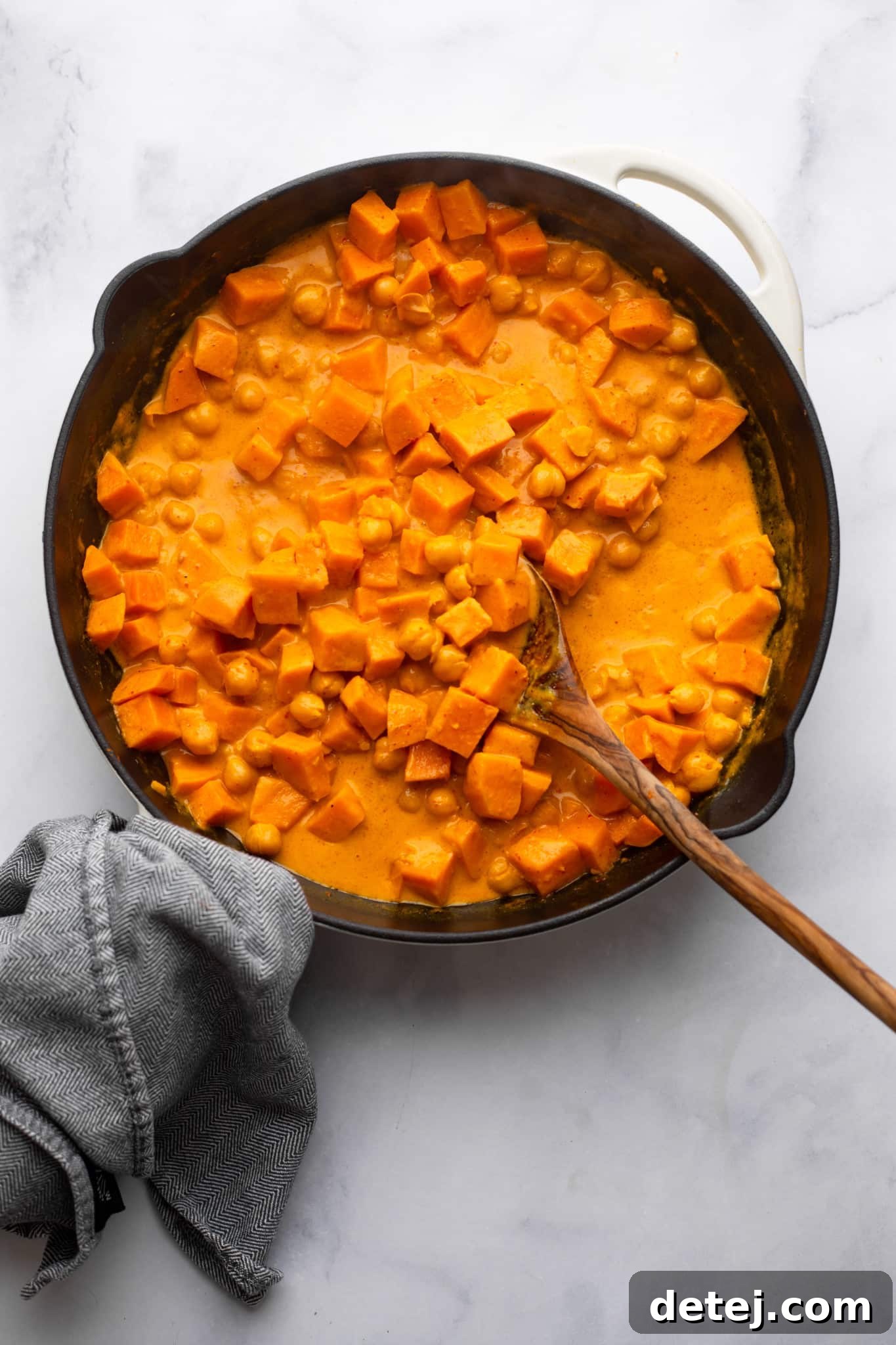 stirring sweet potatoes into a red curry sauce with a wooden spoon in a black skillet.