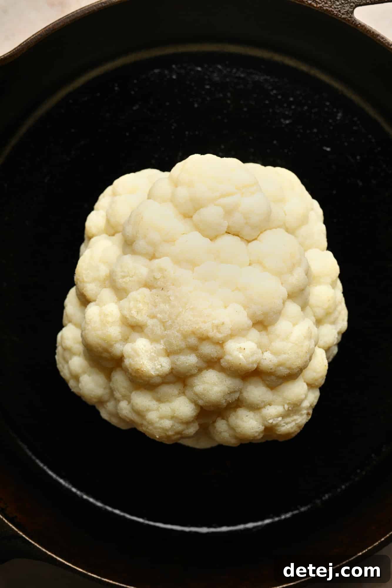 A whole cauliflower head, perfectly boiled and ready for glazing, sits upright in a black cast iron skillet, awaiting the next step.