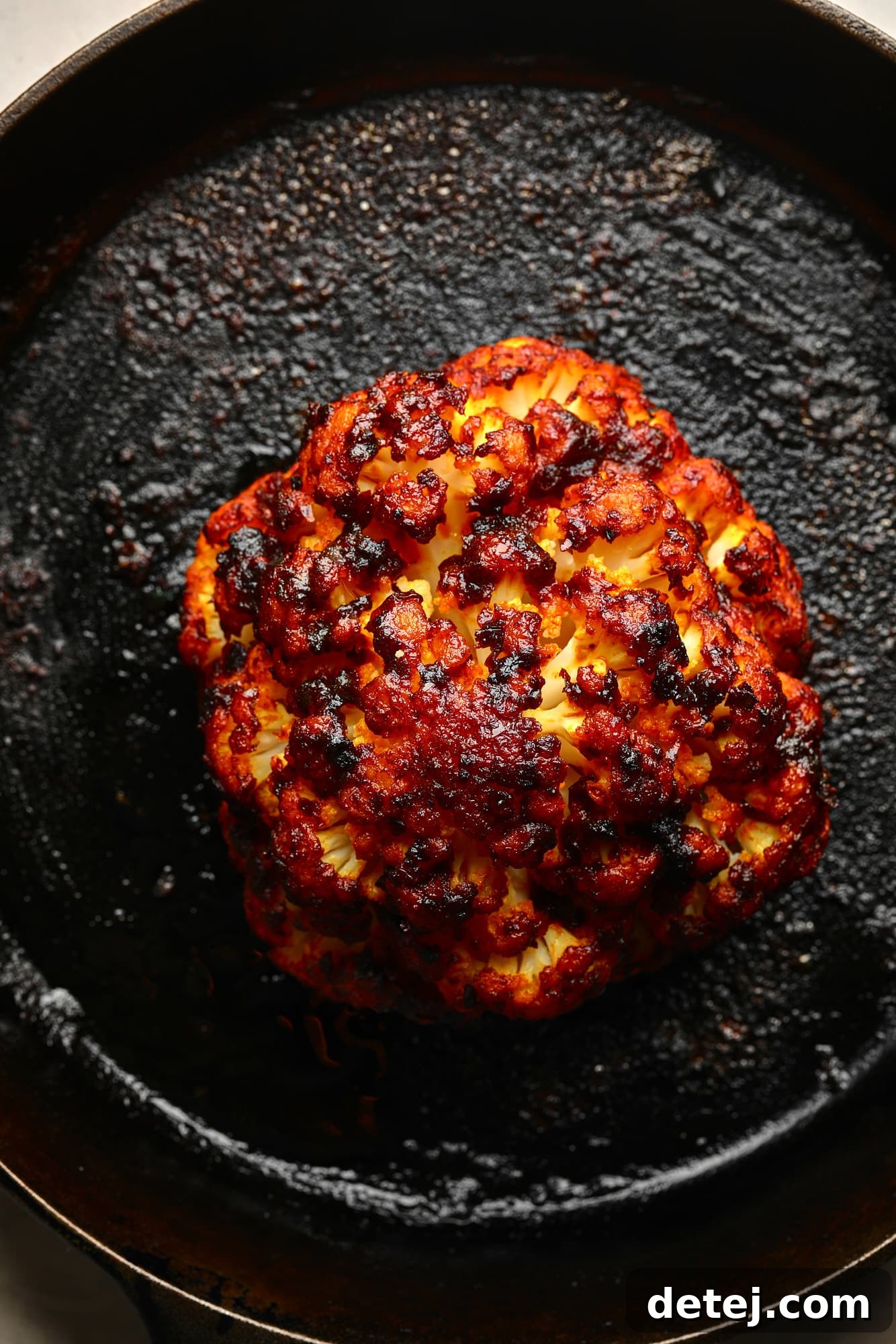 A whole harissa-glazed roasted cauliflower head, beautifully charred and glistening, resting elegantly in a black cast iron skillet, ready to be served.