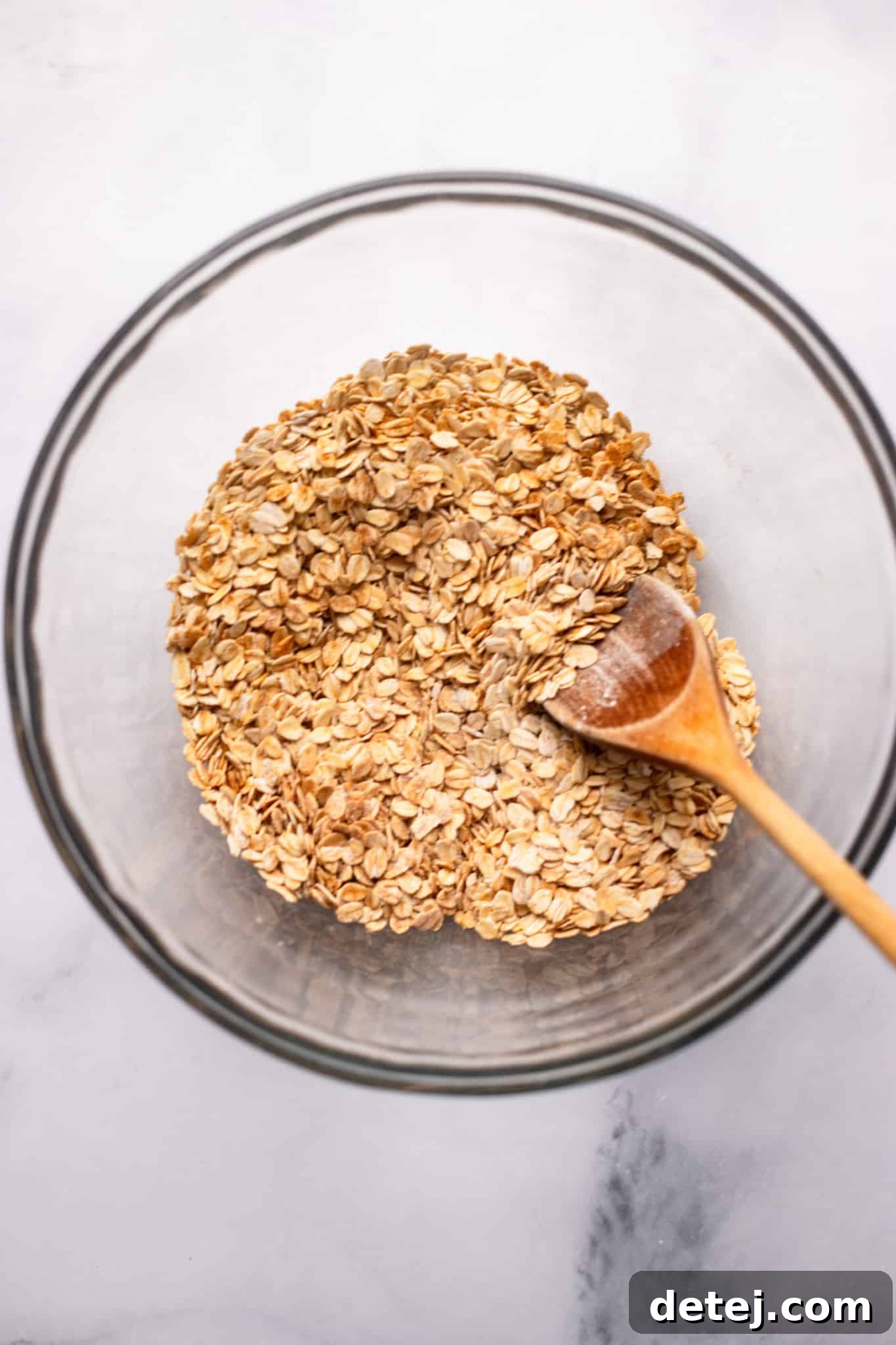 Warm Apple Oat Bake 4 A wooden spoon stirring rolled oats in a clear glass bowl, showing the initial mixing of dry ingredients for baked oatmeal.