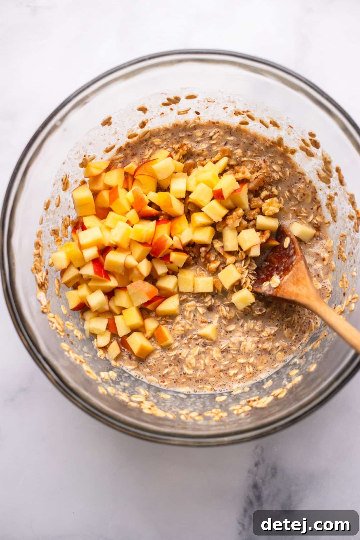 Warm Apple Oat Bake 6 A wooden spoon carefully folding chopped apples into a creamy oatmeal mixture in a glass bowl.