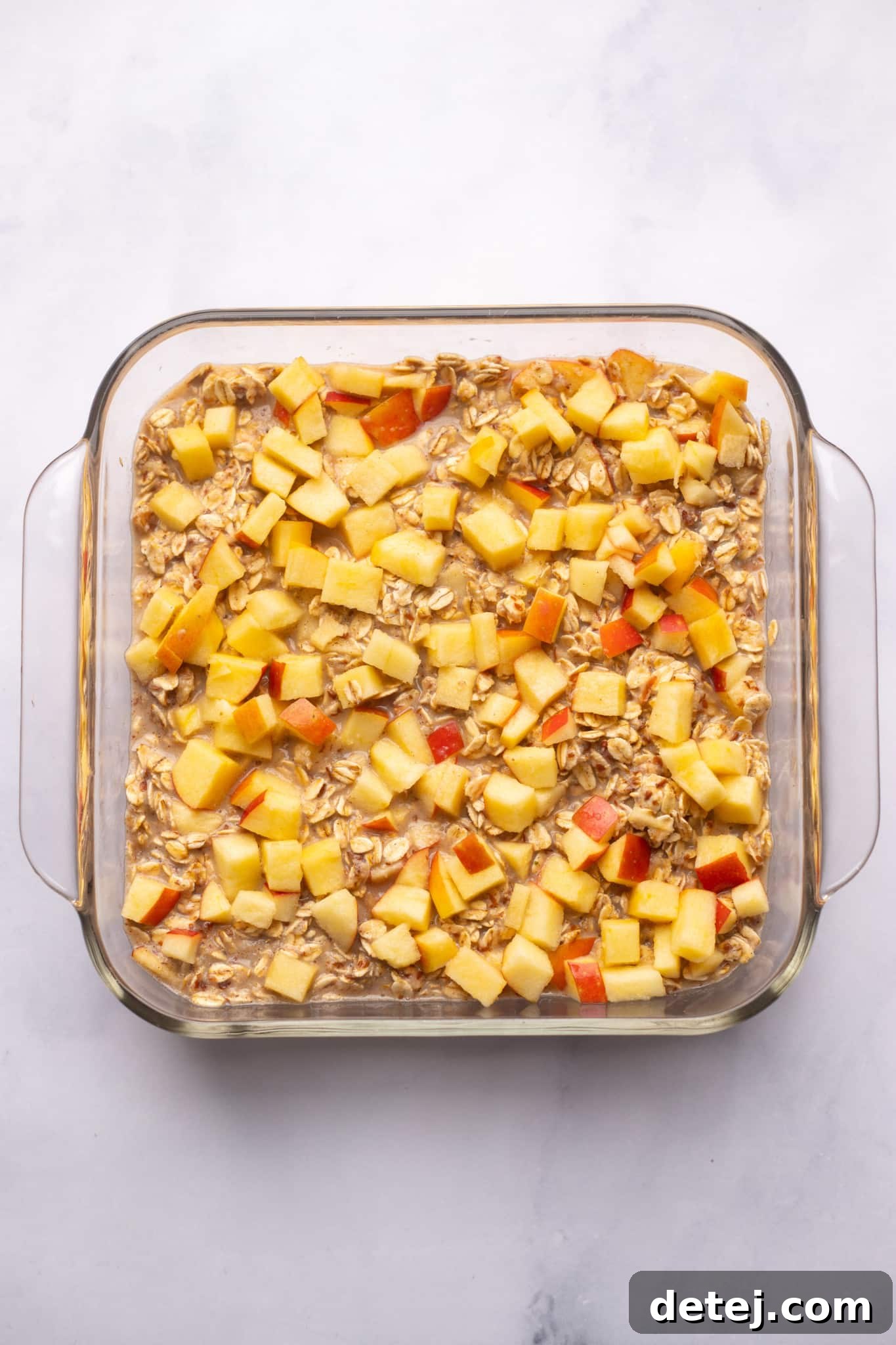 Warm Apple Oat Bake 7 An unbaked apple baked oatmeal mixture in a clear glass baking dish, topped with diced apples, ready for the oven.