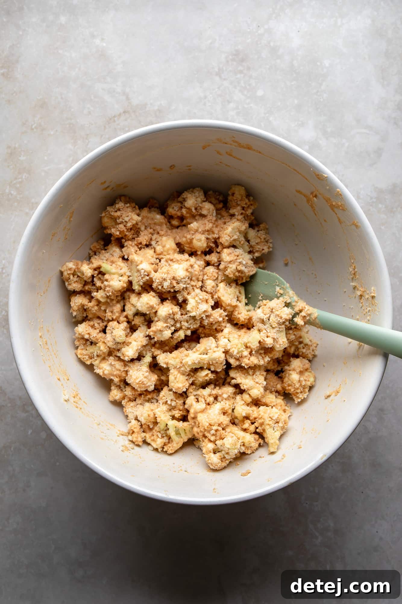 A spatula carefully coating cauliflower florets in a seasoned batter within a white mixing bowl.