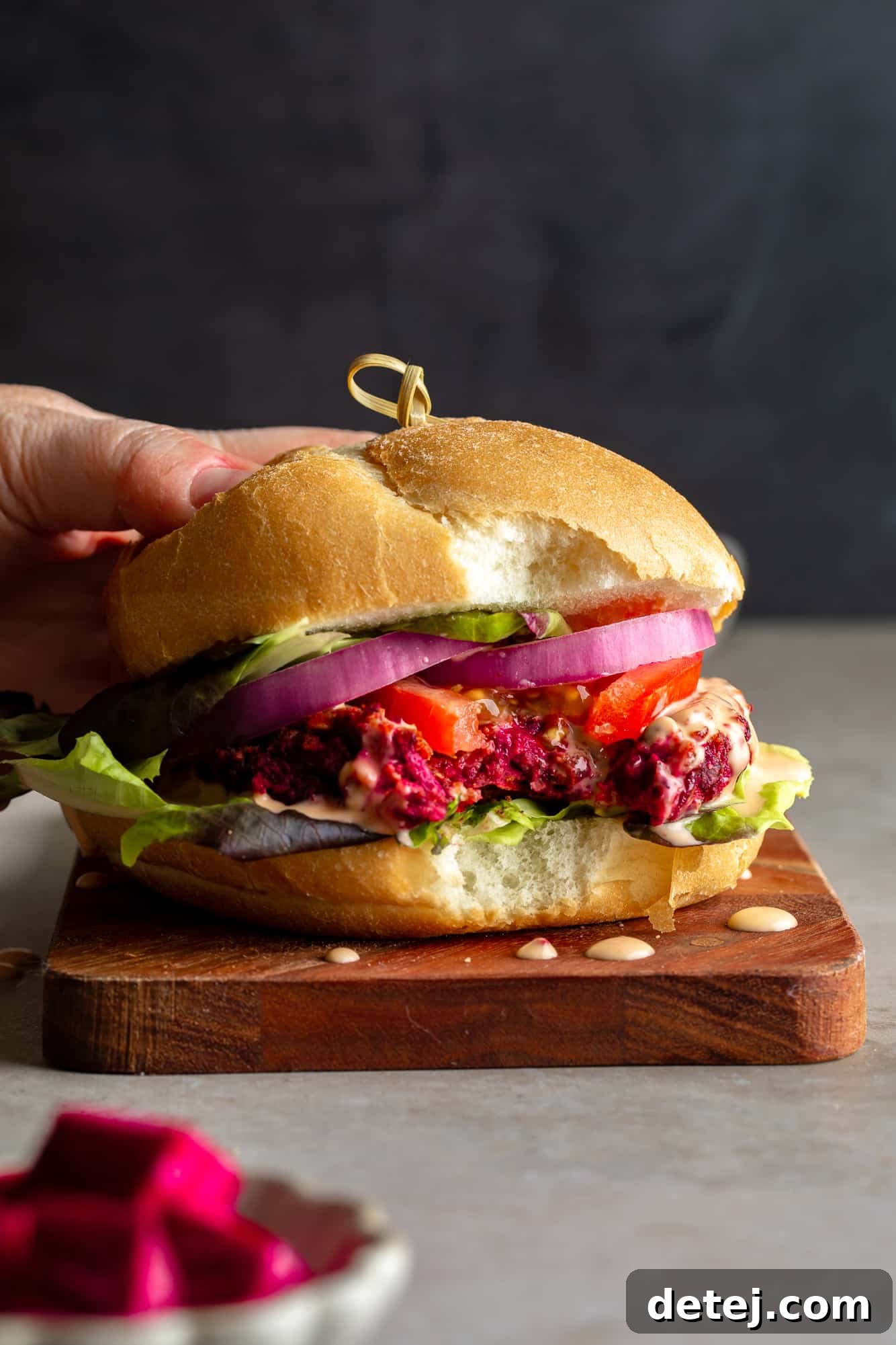 Ruby Red Bean Burgers 2 A woman's hand holding a vibrant kidney beet burger with a bite taken out of it, showcasing its tender, meaty interior and beautiful red color. Garnished with fresh toppings and a creamy sauce.