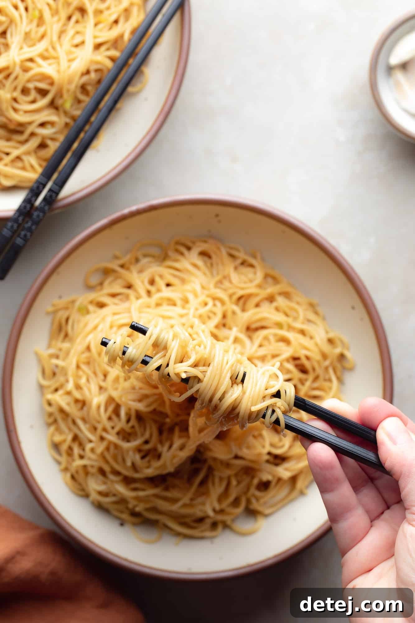 A hand lifting buttery garlic noodles with chopsticks from a white bowl, showcasing their delicious texture.