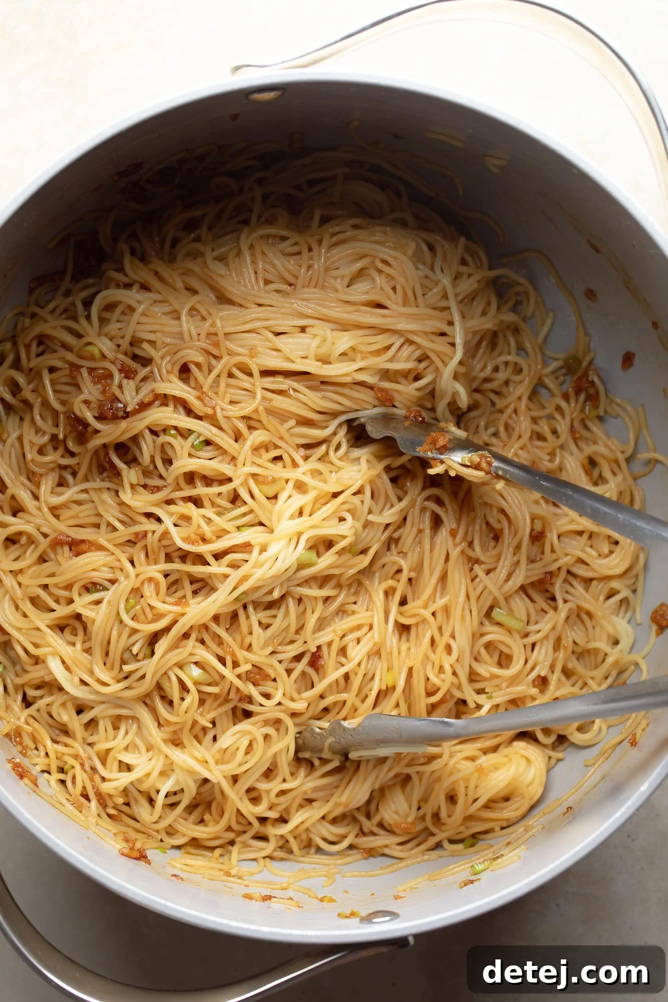 Tongs gently tossing cooked noodles with a luscious garlic sauce in a skillet, ensuring every strand is coated.
