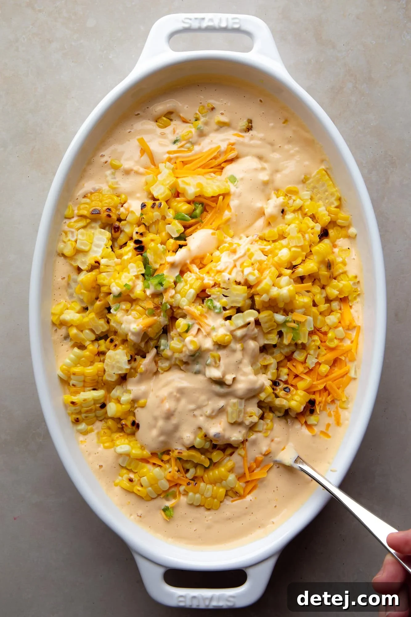 A spoon meticulously combines the creamy cashew sauce, charred corn, vegan cheese, and jalapeño in a baking dish, ready for baking.