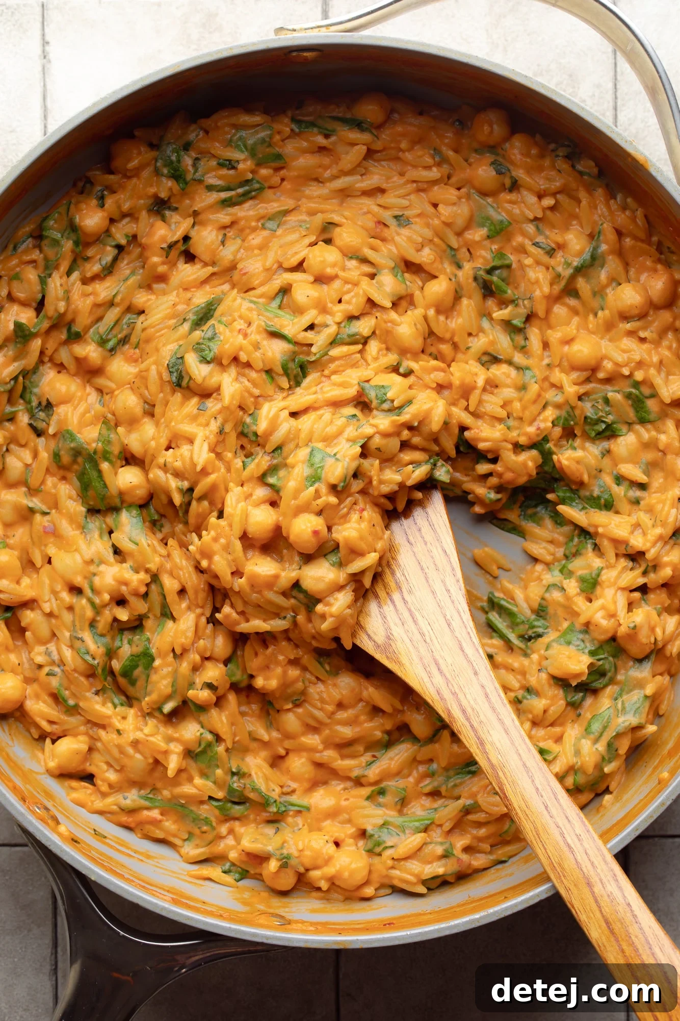 A wooden spoon stirring the creamy tomato orzo and chickpeas in a large skillet, showing the rich texture.