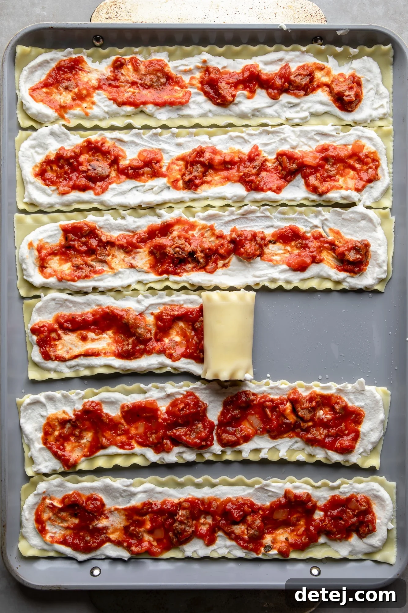 The process of rolling up cheese and sauce-layered lasagna noodles on a baking sheet, illustrating the assembly step of vegan lasagna rolls.