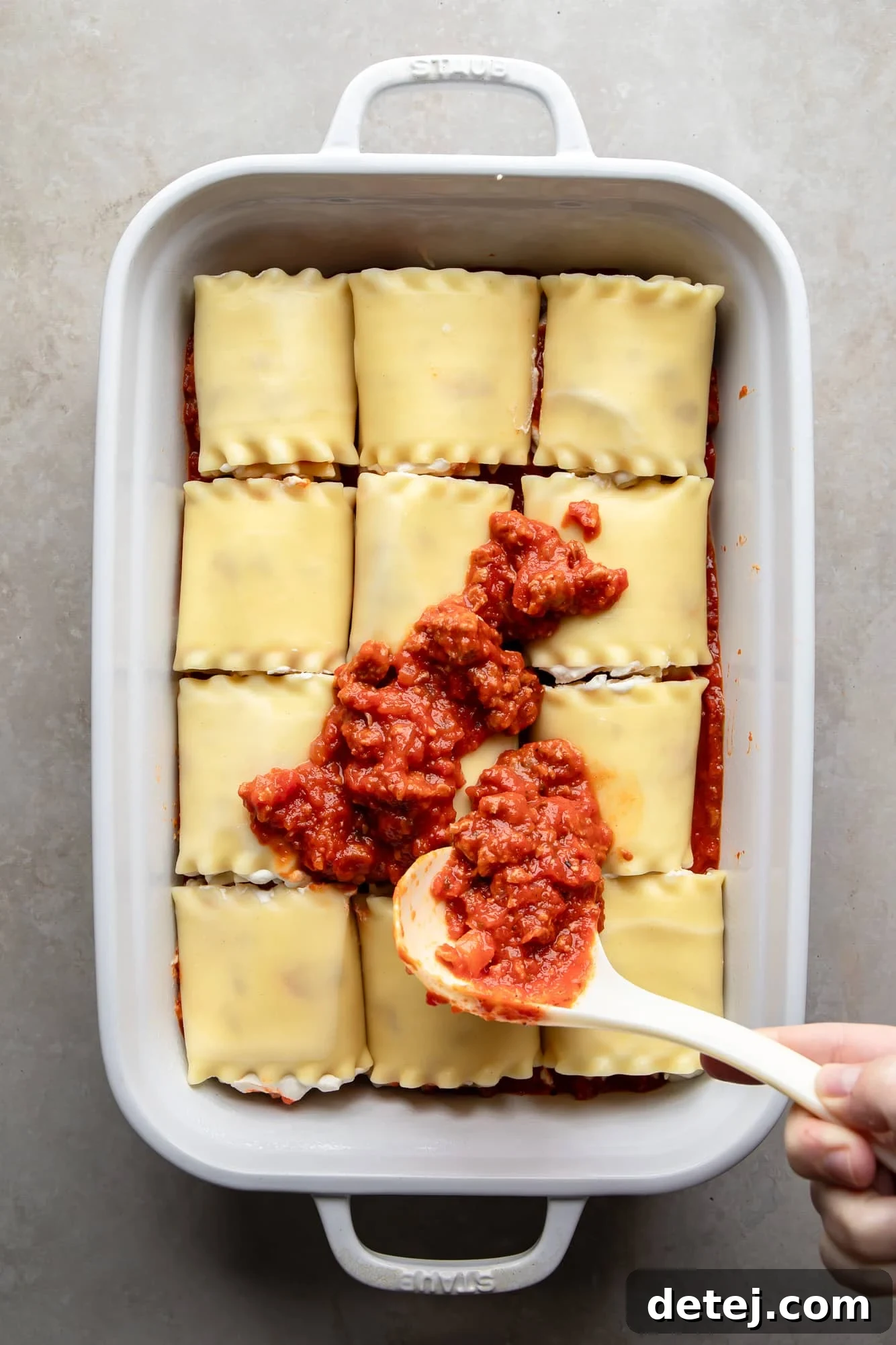 Spoonfuls of rich marinara sauce being spread over assembled vegan lasagna rolls in a white casserole dish, preparing them for baking.