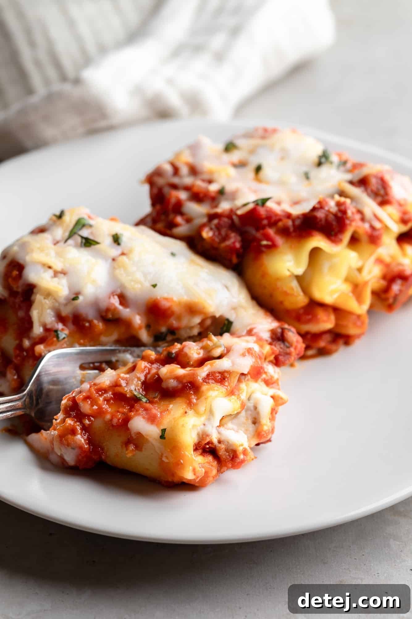 Using a fork to cut into a perfectly baked vegan lasagna roll on a white plate, revealing its creamy layers and savory sauce.