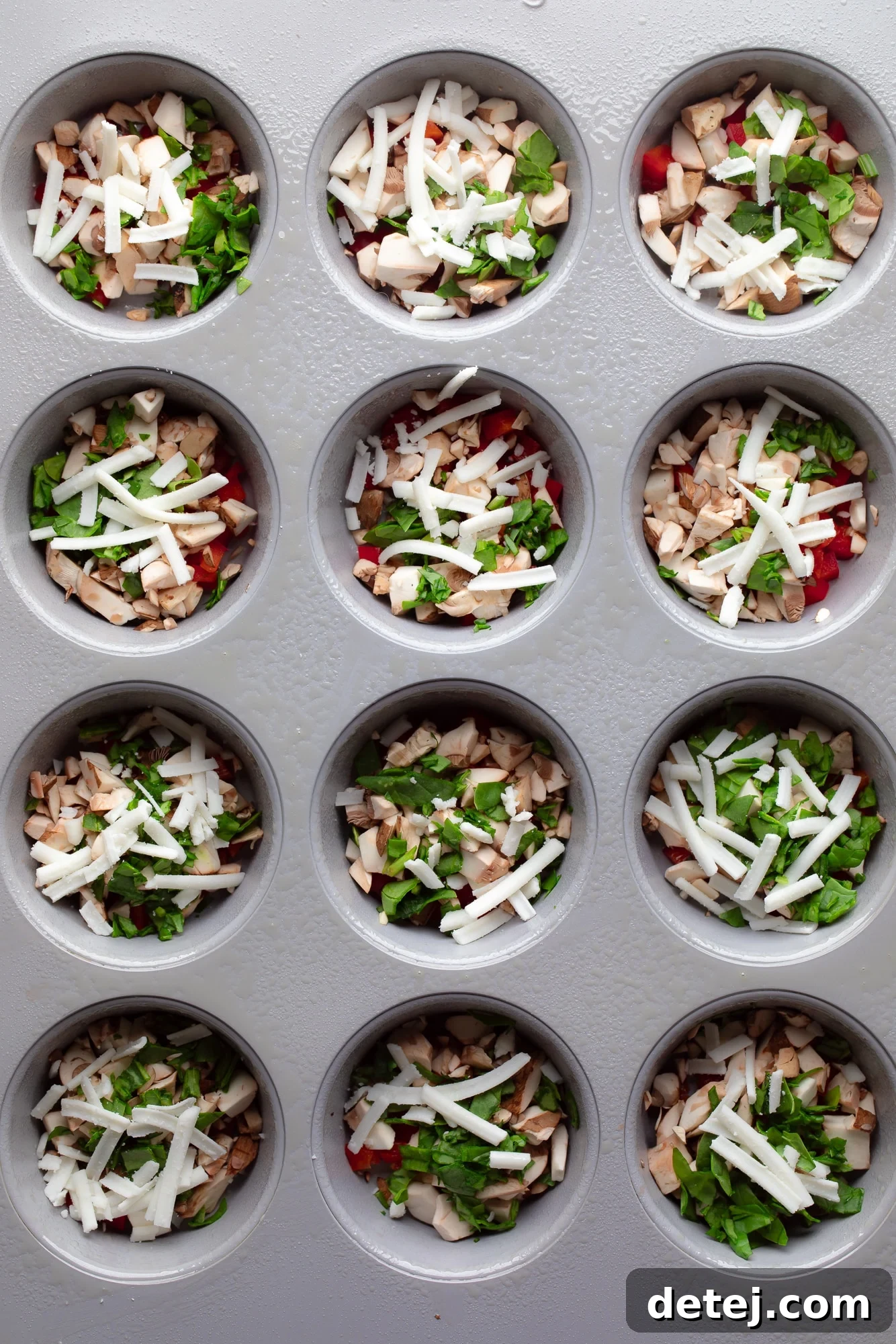 A muffin tin with individual cups filled with a colorful mix of diced red bell peppers, chopped mushrooms, fresh spinach, and shredded vegan cheese, ready for the liquid egg mixture.