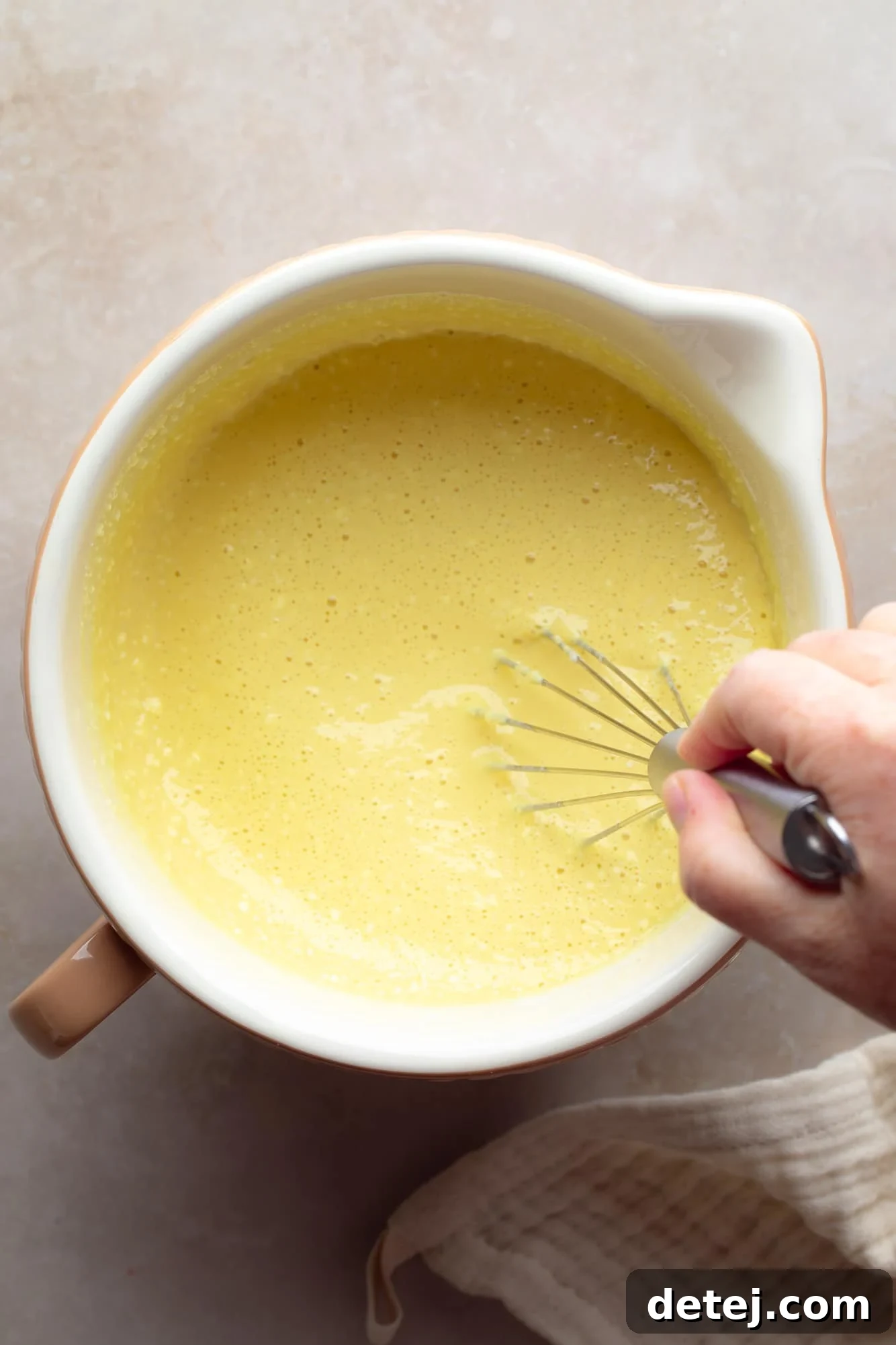 Close-up of a glass bowl containing the smooth, pale yellow liquid JUST Egg mixture, being whisked with a metal whisk, ready to be poured into the muffin cups.