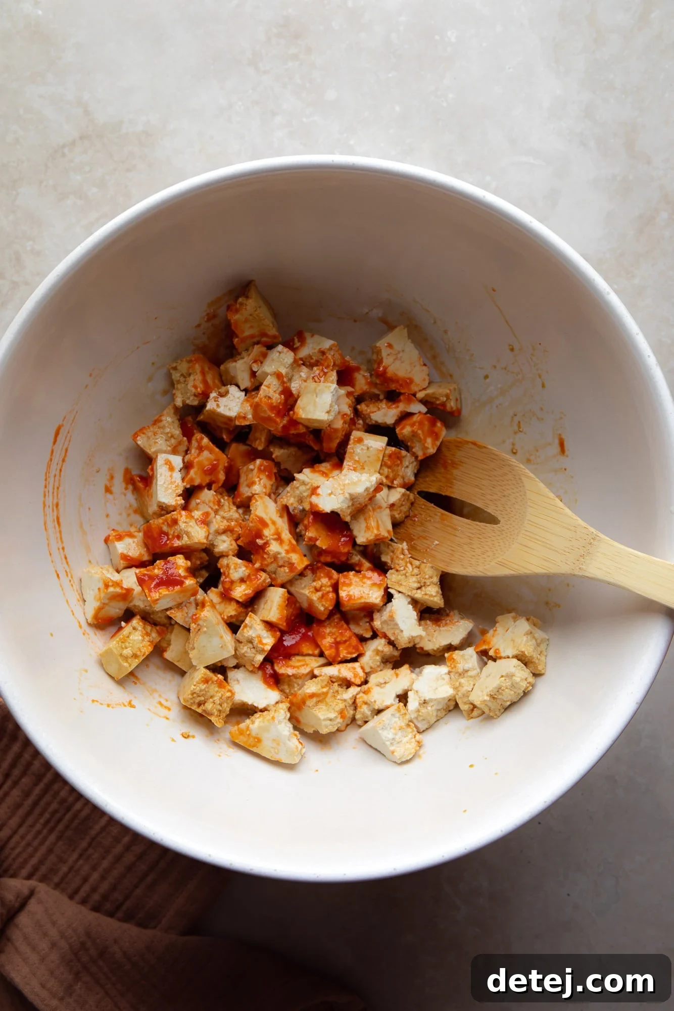 using a wooden spoon to toss tofu cubes in hot sauce and seasonings in a large white bowl.