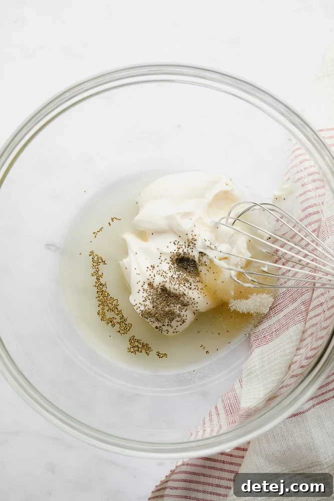 A clear glass bowl containing vegan mayonnaise, sugar, apple cider vinegar, and celery seed, before mixing.