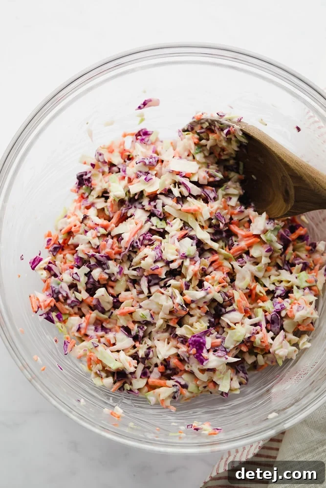A wooden spoon mixing creamy dressing and shredded cabbage in a glass bowl.