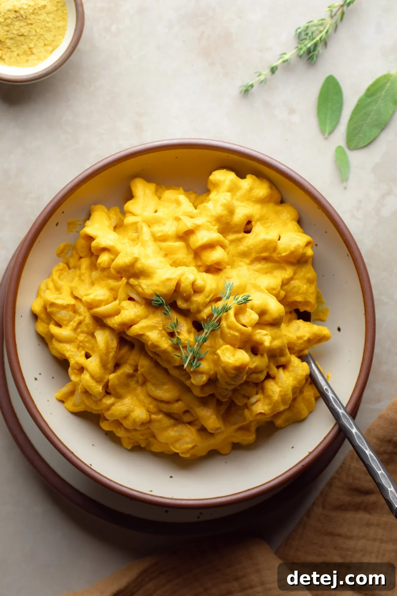 A vibrant bowl of vegan pumpkin mac and cheese, garnished with a sprig of fresh thyme, photographed from an overhead perspective. The pasta is coated in a rich, creamy orange sauce, inviting warmth and comfort.