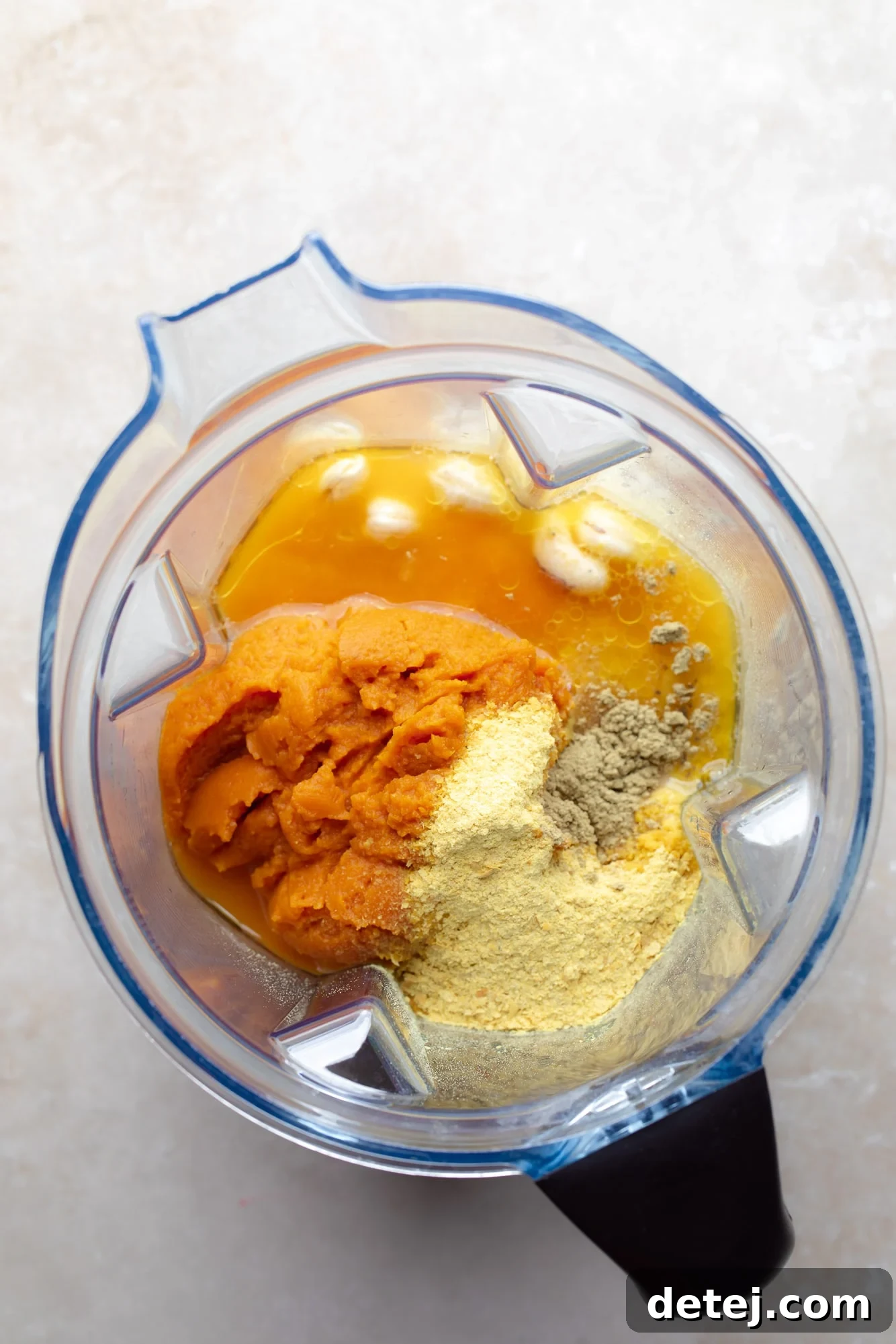 Raw ingredients for vegan pumpkin mac and cheese, including soaked cashews, pumpkin puree, nutritional yeast, and sautéed onions and garlic, prepared for blending in a high-powered blender cup.