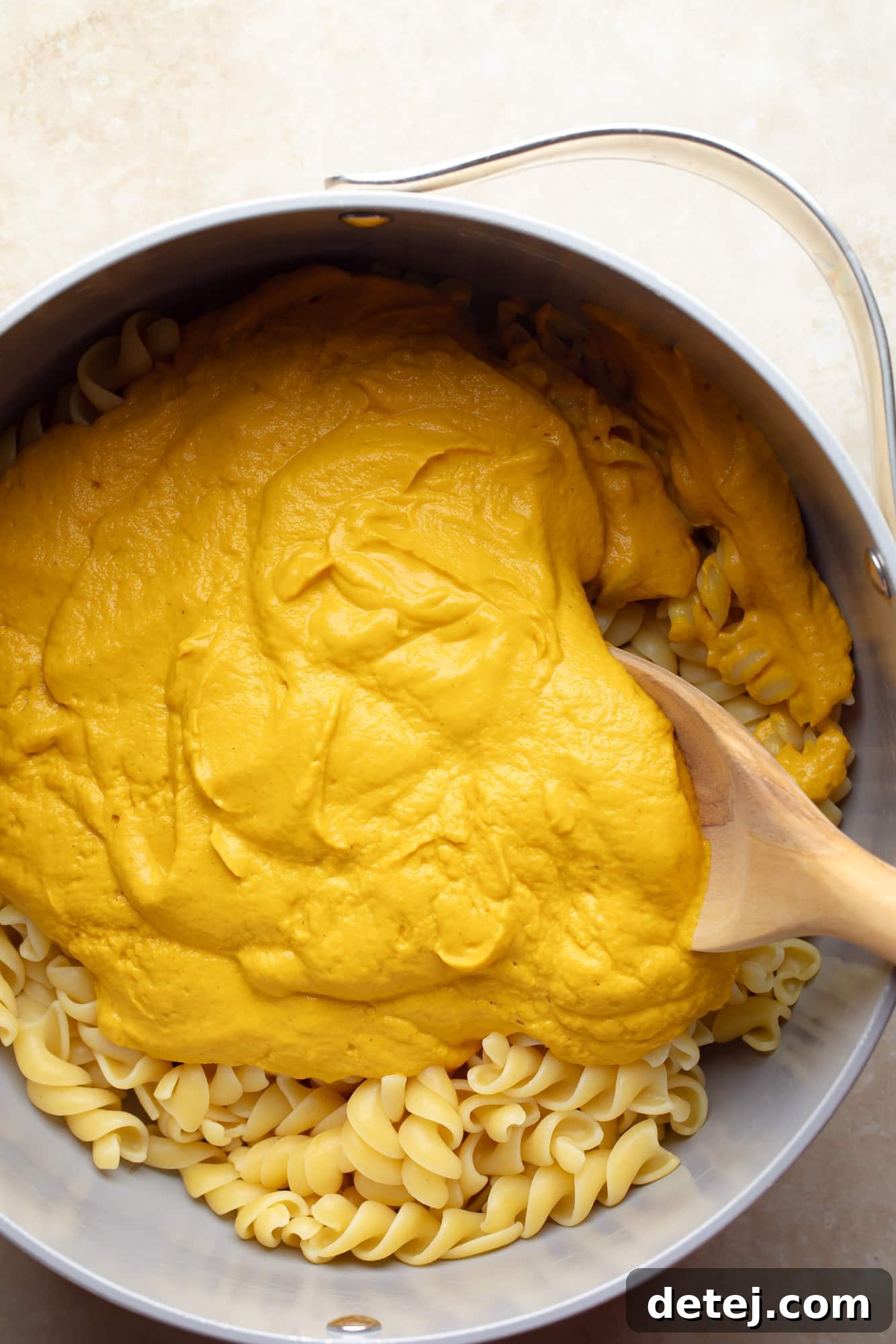 A wooden spoon stirring pasta noodles coated in a luscious, orange vegan pumpkin cheese sauce within a cooking pot. The steam indicates warmth and fresh preparation, showcasing the perfect consistency of the sauce.