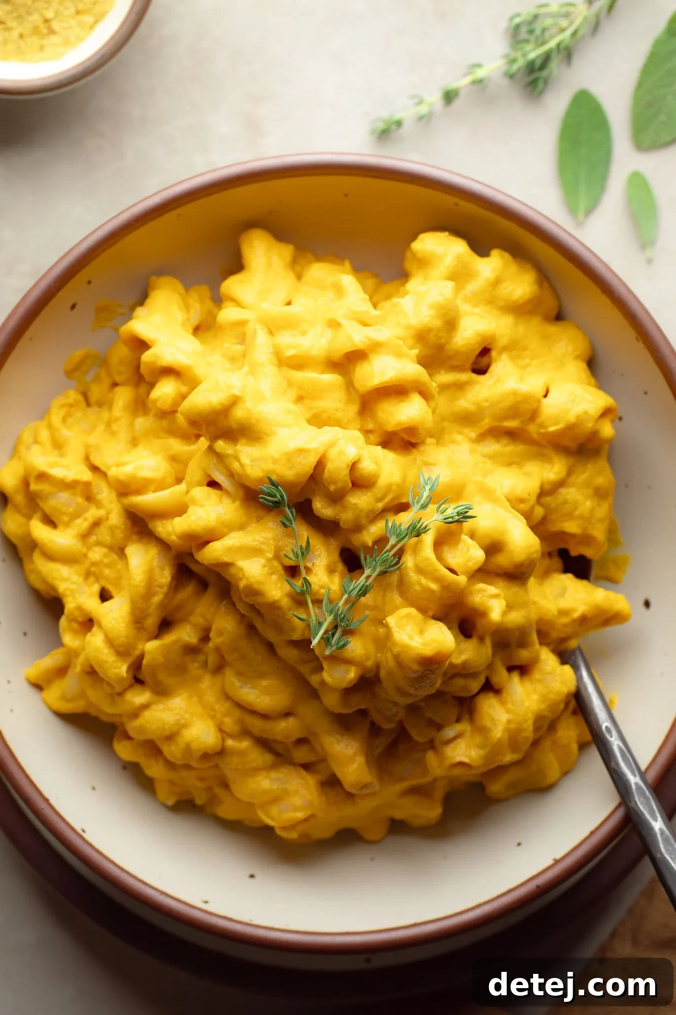 A high-angle view of a bowl of creamy vegan pumpkin mac and cheese, elegantly garnished with a fresh sprig of thyme. The presentation is inviting, highlighting the rich texture and vibrant color of the dish.