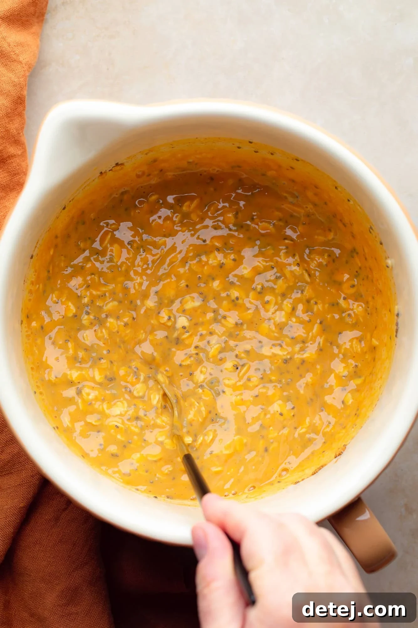 a womans hand stirring a pumpkin overnight oats mixture in a large white bowl.