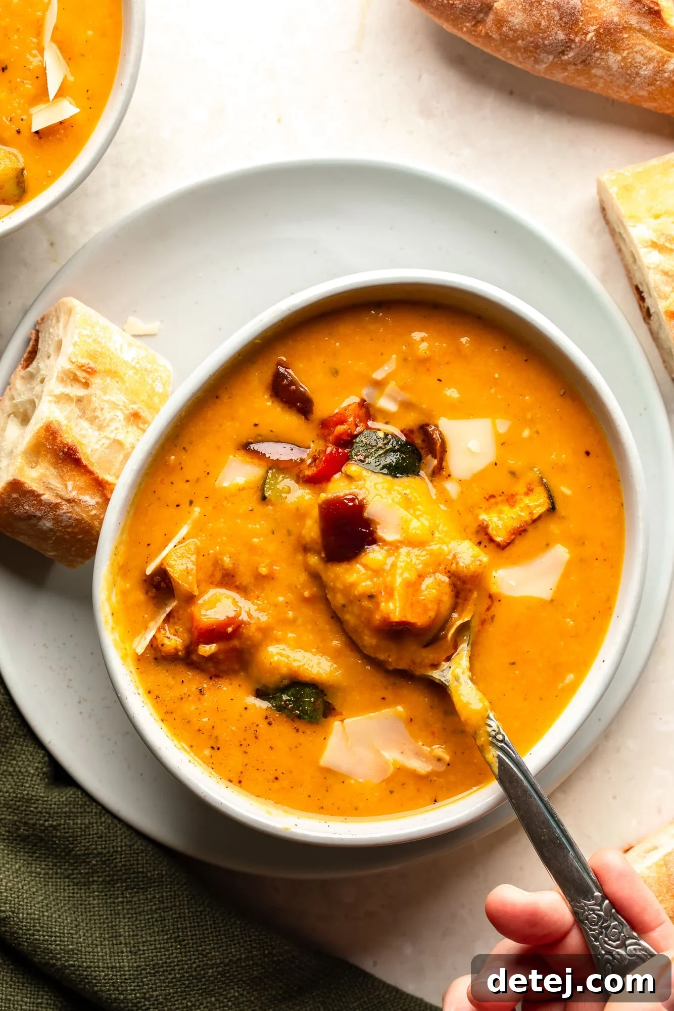 Overhead view of a woman's hand lifting a spoon out of a bowl of creamy roasted vegetable soup, showing the rich texture.