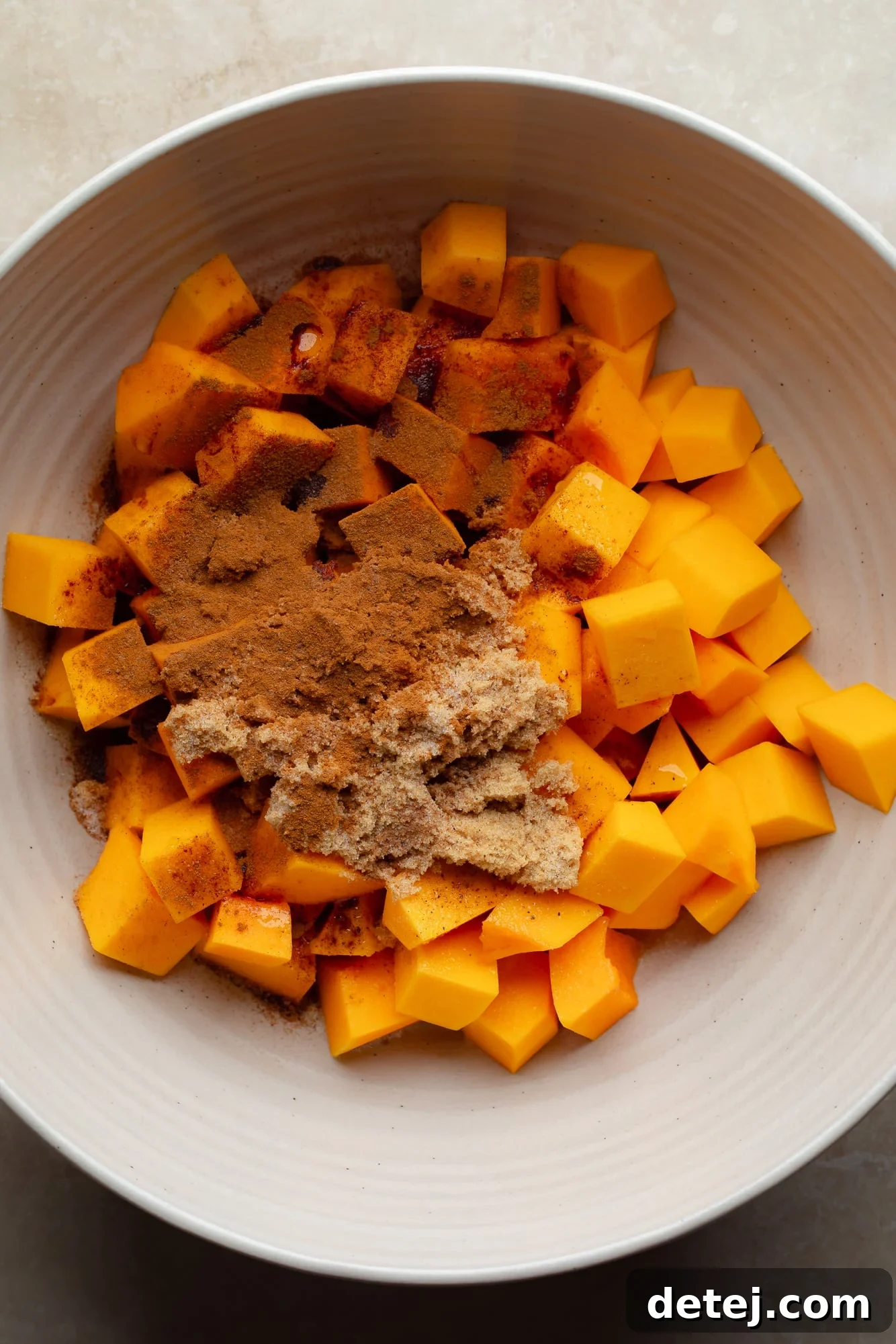 overhead view of spices sprinkled on top of raw butternut squash cubes in a large bowl.
