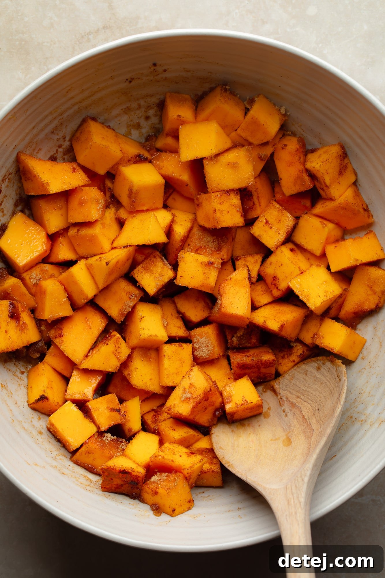 using a wooden spoon to toss raw butternut squash cubes in spices in a large bowl.