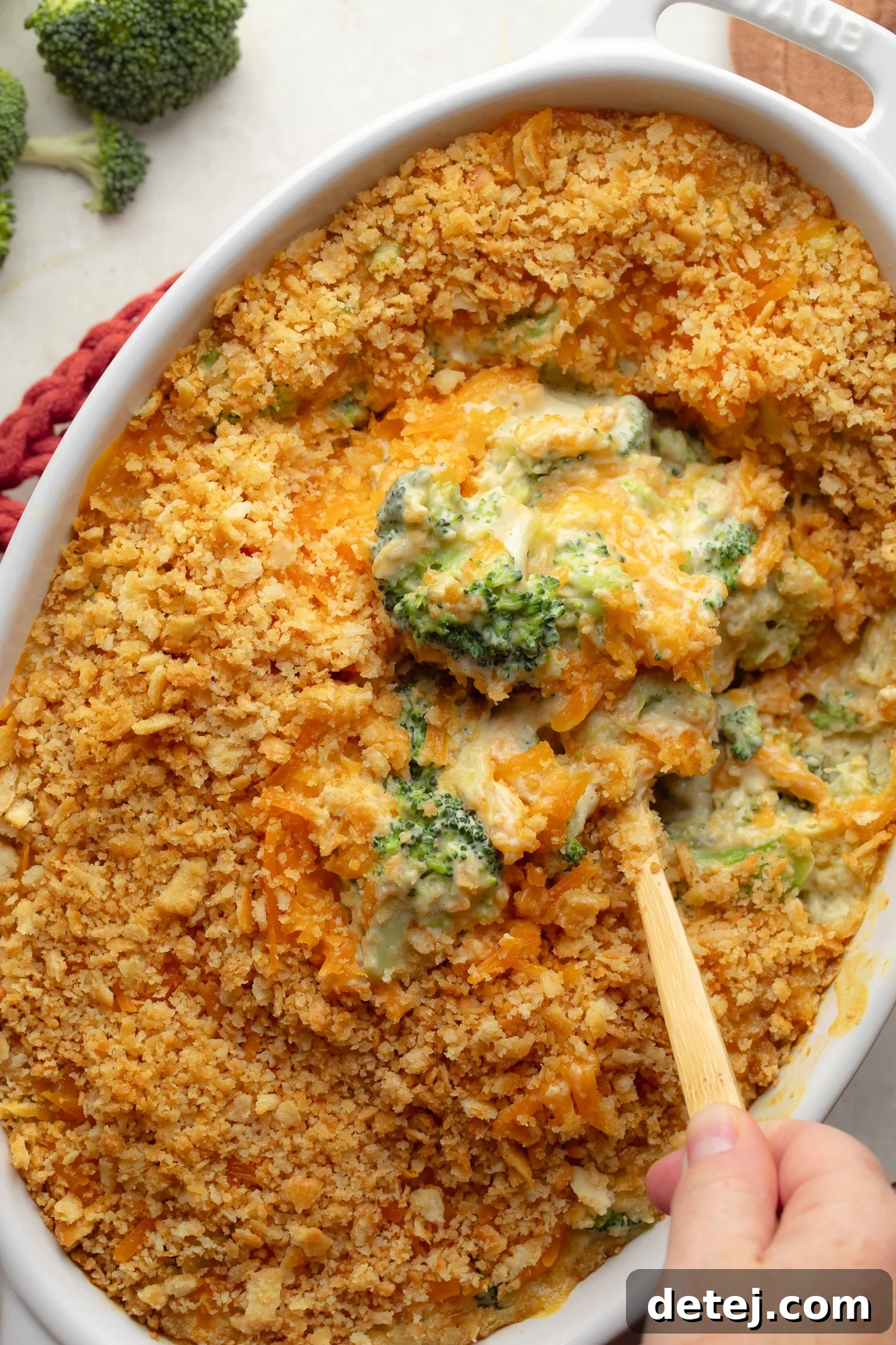 A hand using a spoon to lift a scoop of rich, creamy vegan broccoli casserole from a large baking dish, showcasing its cheesy texture and crispy topping.