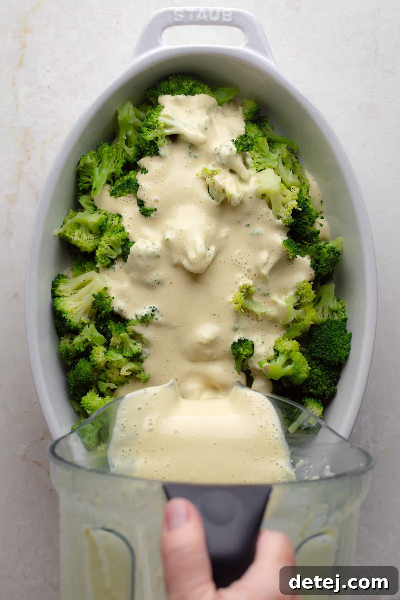 A woman's hand gracefully pouring a luscious vegan cheese sauce over a bed of vibrant green broccoli florets in a large casserole dish, ready for tossing.