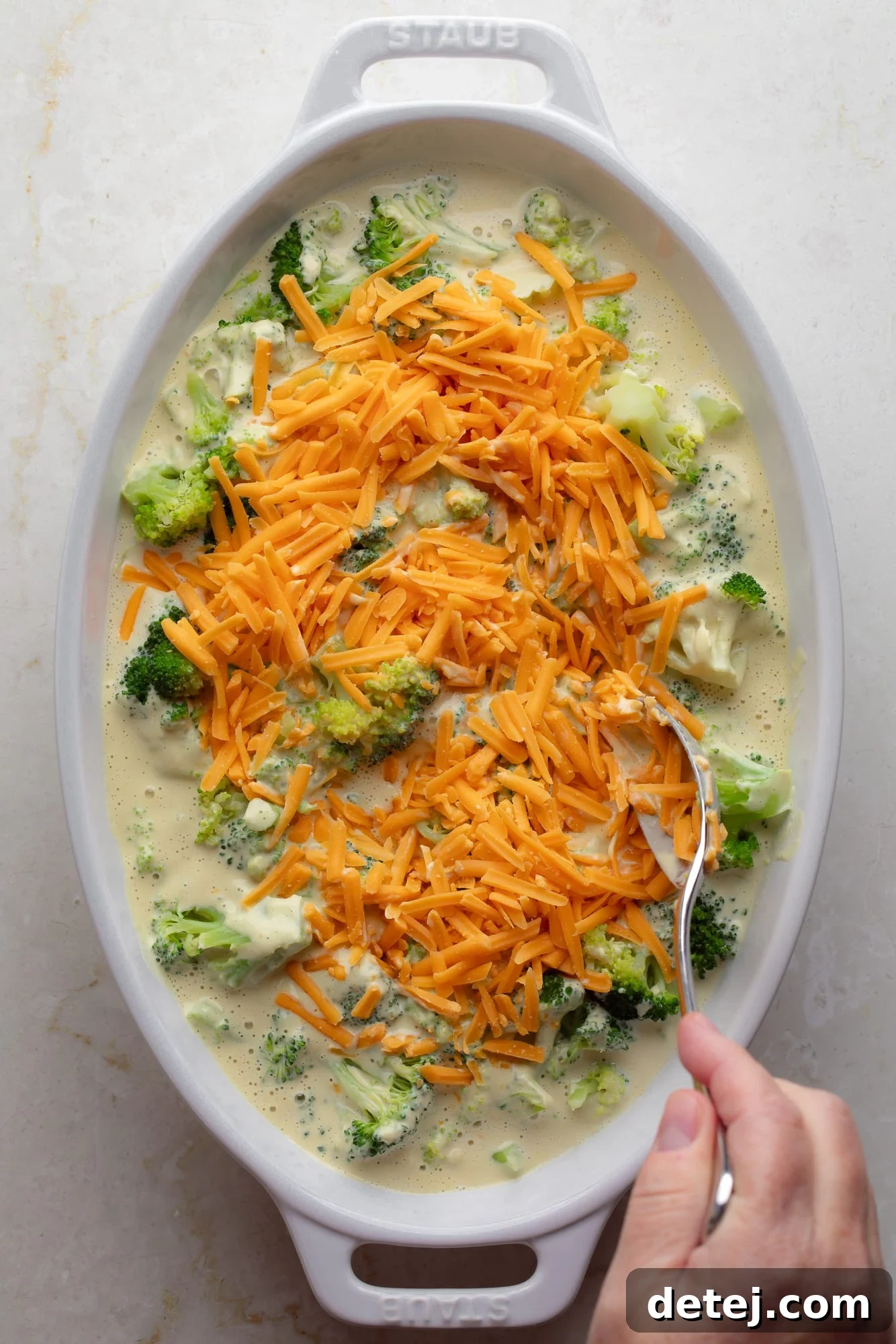 A woman's hand stirring gooey vegan cheese shreds into the creamy vegan broccoli casserole mixture in a large dish, illustrating the assembly process.