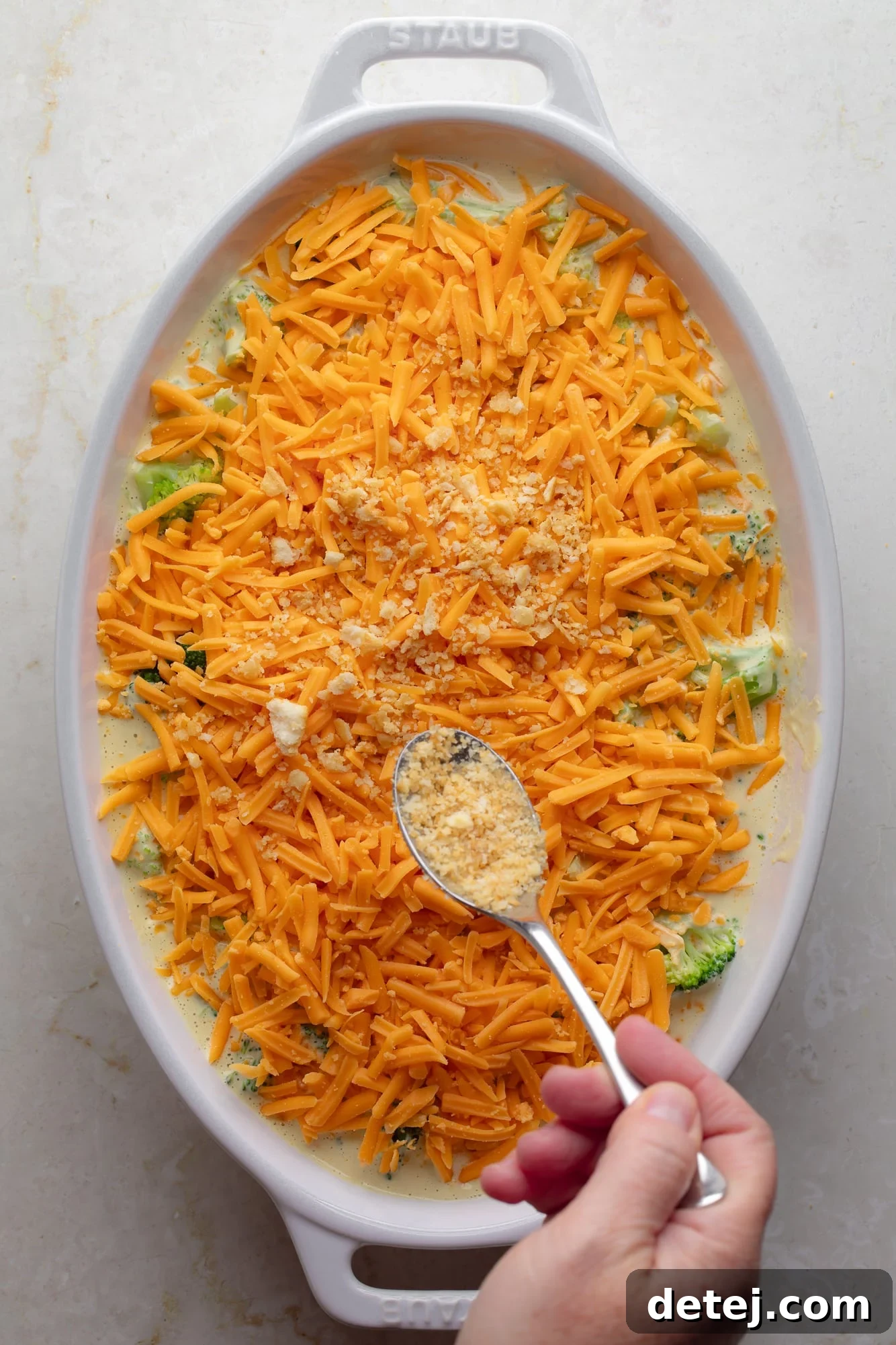 A woman's hand delicately sprinkling golden, buttery breadcrumbs over a vegan cheese-topped vegan broccoli casserole, preparing it for baking.