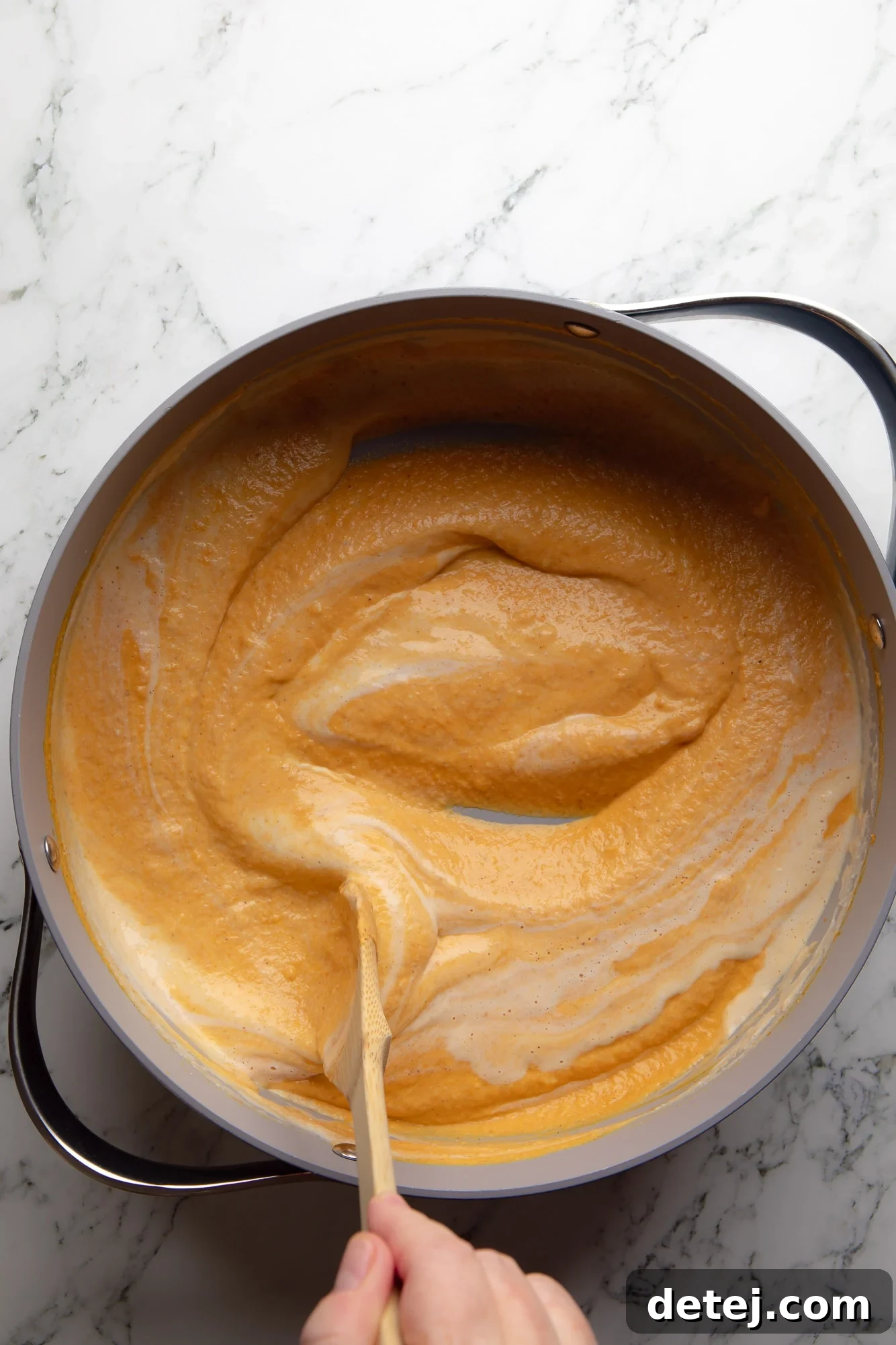 Overhead shot of a hand stirring rich pumpkin sauce with a wooden spoon in a skillet, showcasing the vibrant orange color.