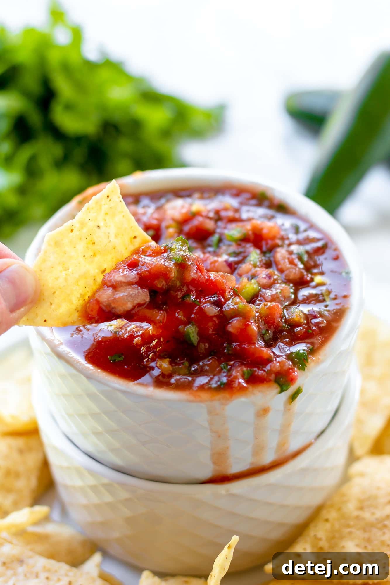 Effortless Fresh Salsa 5 Woman's hand dipping a tortilla chip into a small ceramic bowl of freshly made tomato salsa, against a rustic background.