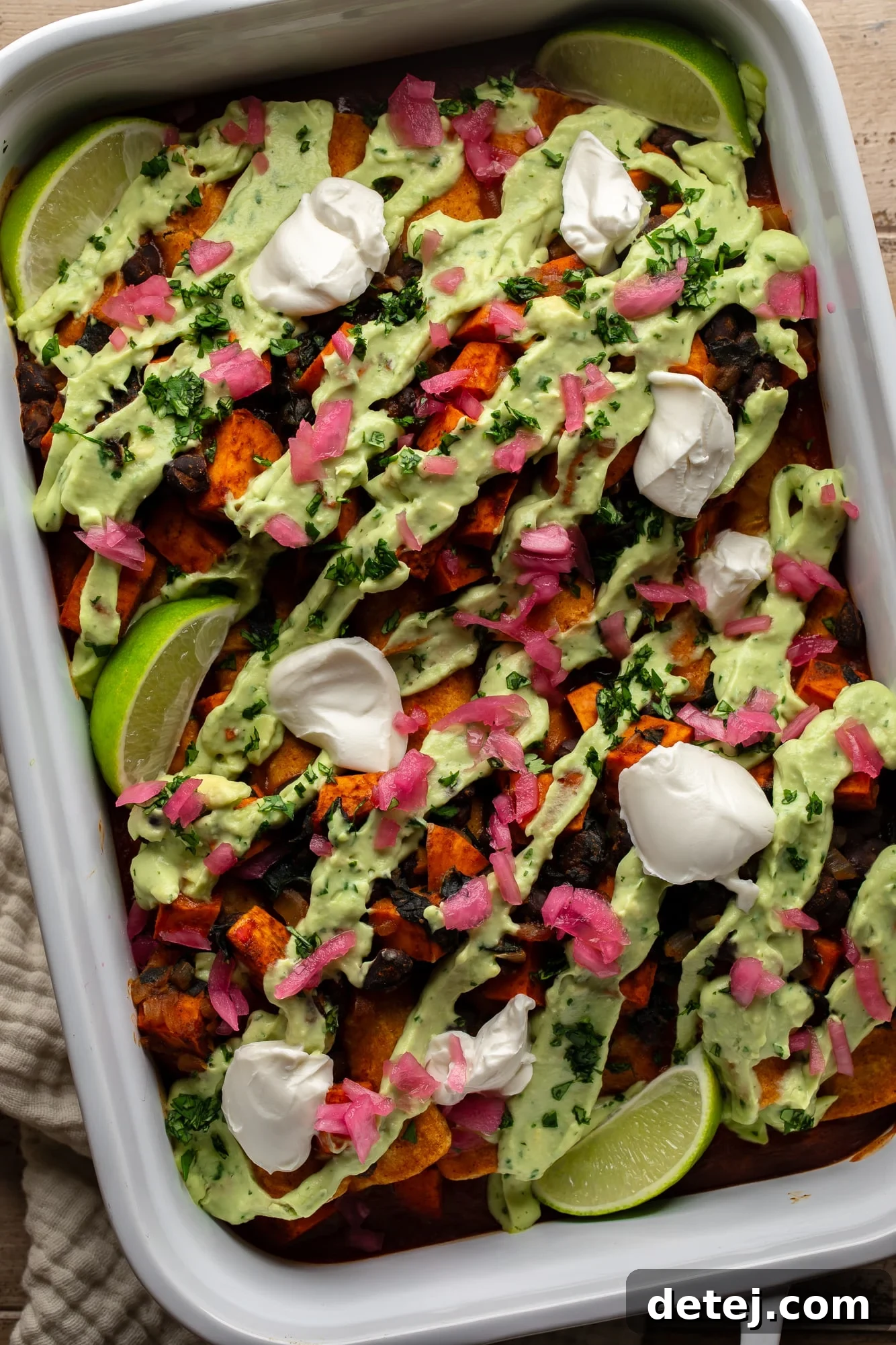 overhead view of sweet potato black bean enchiladas topped with avocado crema, vegan sour cream, pickled red onions, and lime wedges, showcasing the rich colors and textures of the dish.