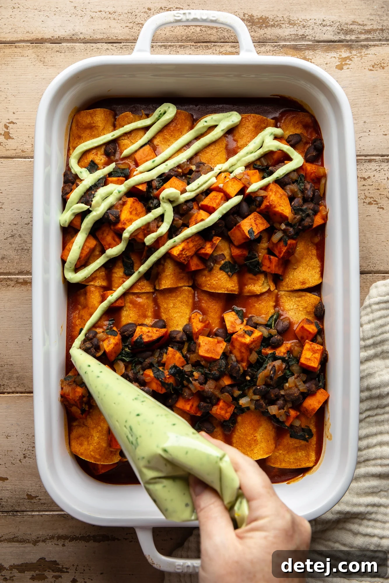 a woman's hand piping vegan avocado crema on top of freshly baked sweet potato black bean enchiladas in a large casserole dish, ready for serving.