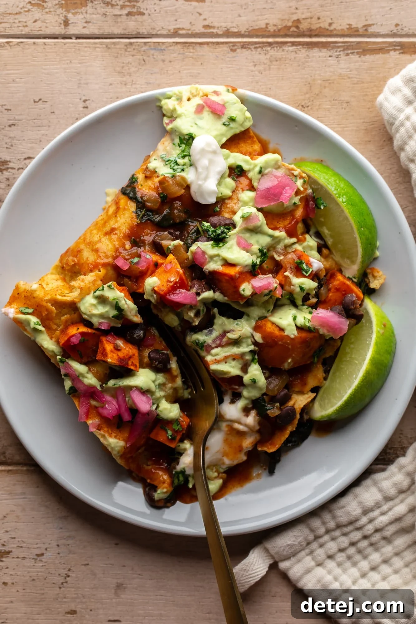 using a fork to cut into sweet potato black bean enchiladas on a plate, showing the creamy avocado crema and vibrant filling.
