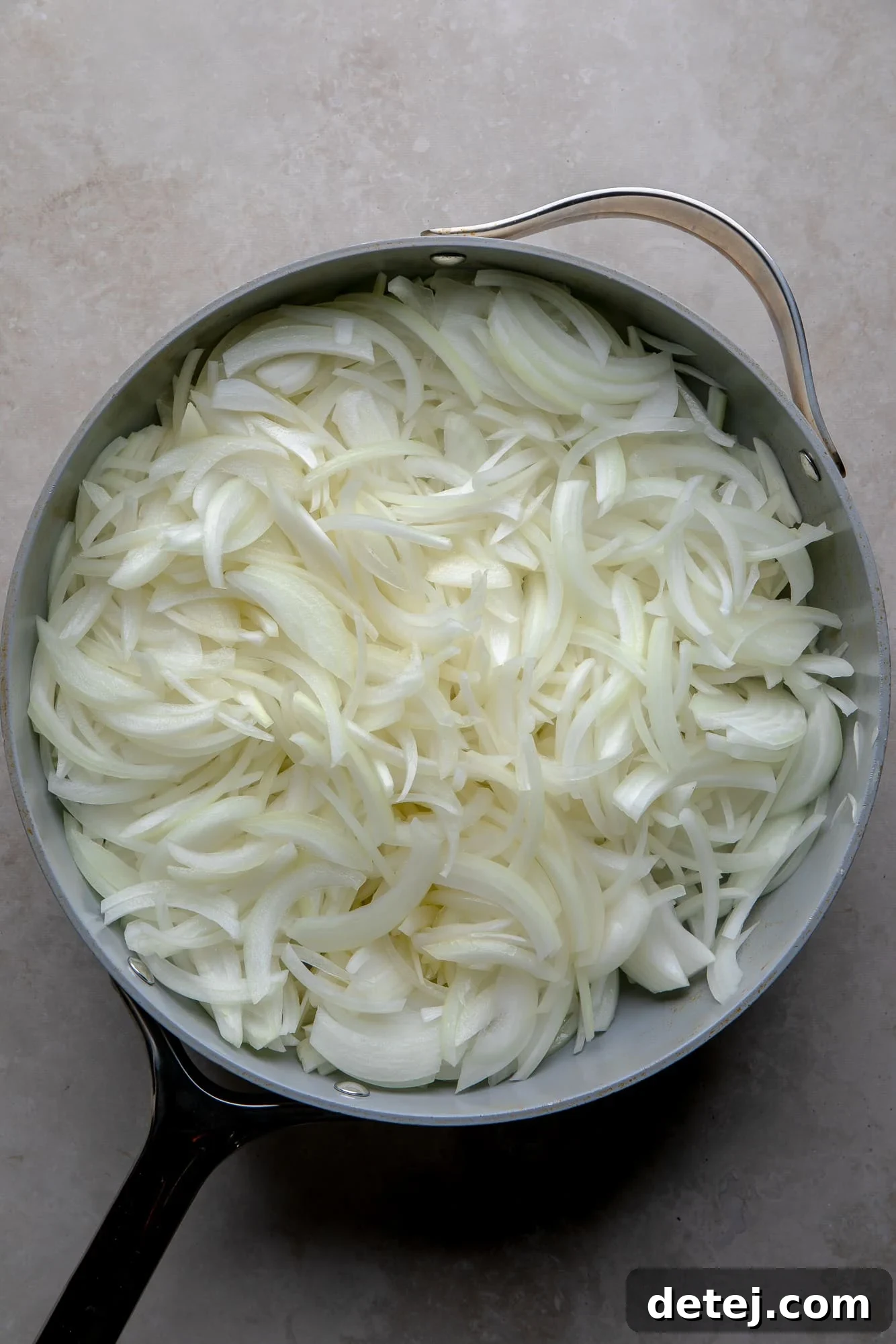 Overhead view of raw, thinly sliced yellow onions starting to cook in a large skillet with olive oil.