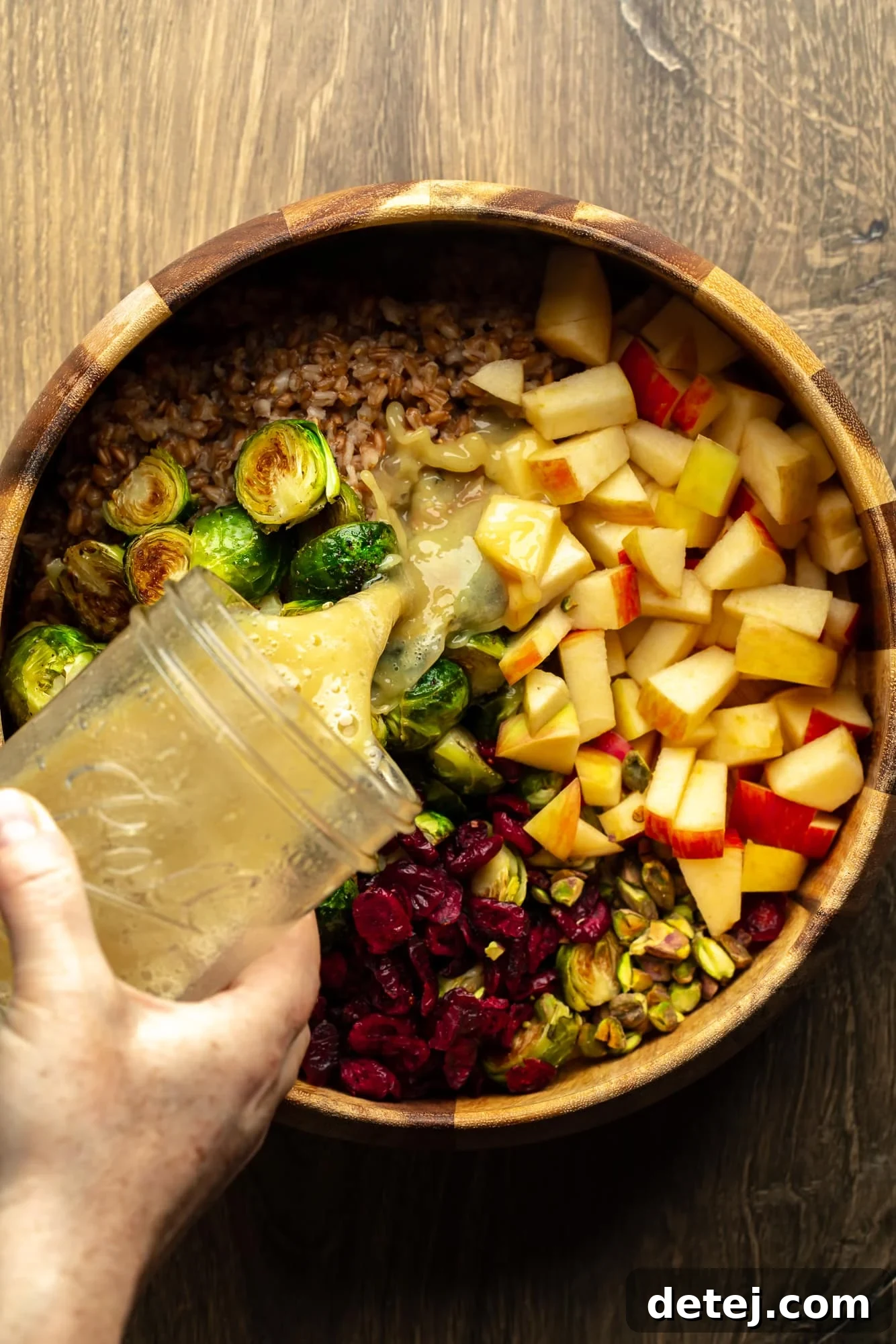 Maple Dijon dressing being drizzled from a mason jar onto a warm brussels sprouts and farro salad in a large mixing bowl, illustrating the final step of flavor infusion.