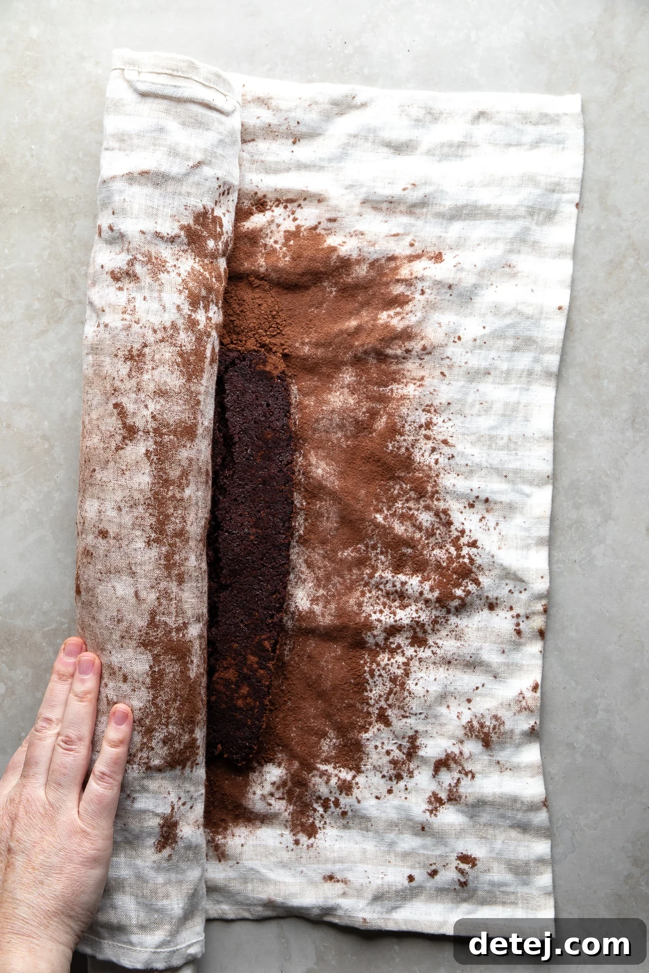 overhead view of a hand rolling a chocolate cake in a white cocoa powder-dusted towel.