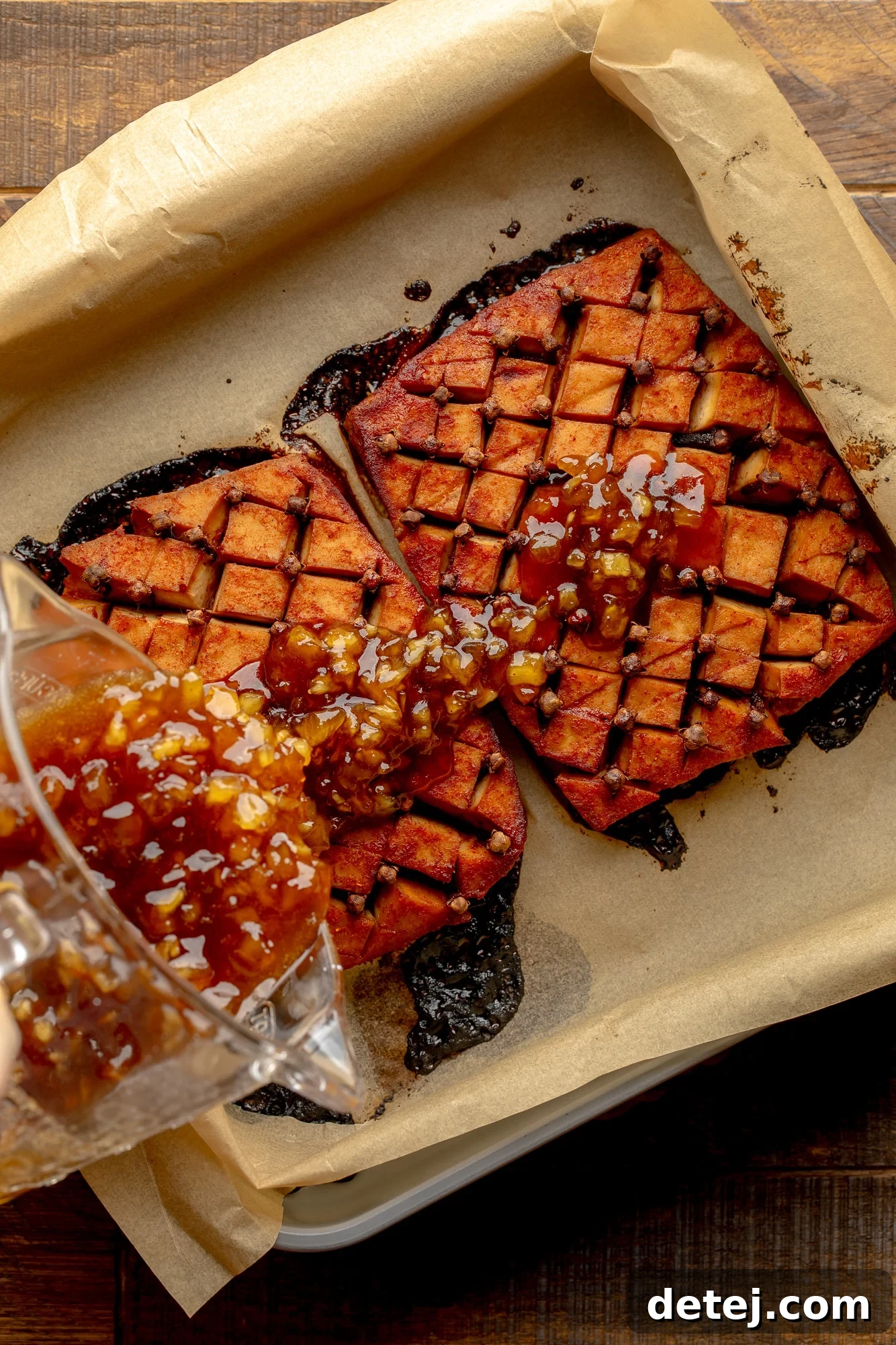 overhead view of a pineapple glaze being poured over a tofu ham roast in a parchment-lined baking dish.